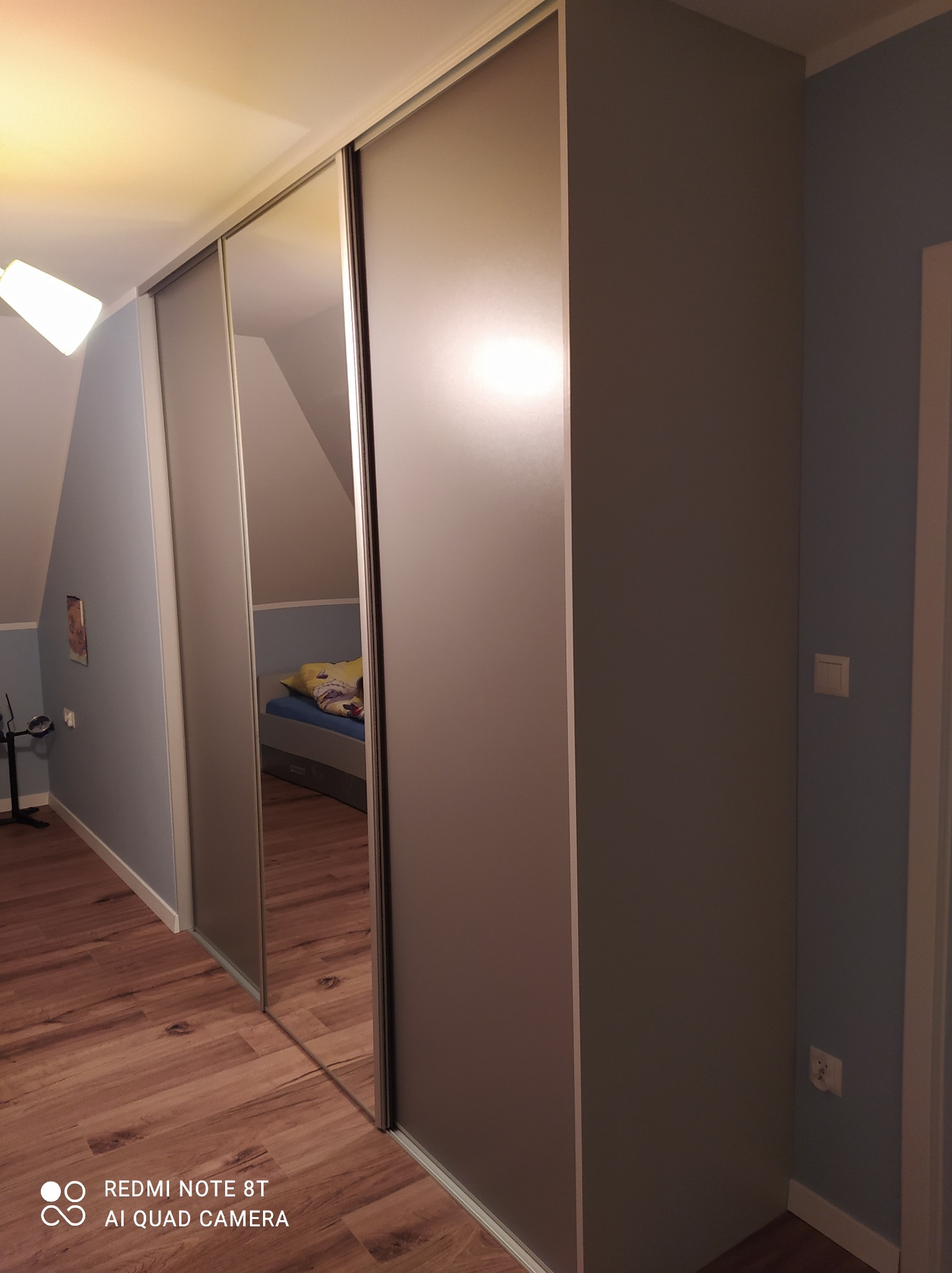 Szafa przesuwna z lustrem w pokoju na poddaszu, widoczne skośne ściany i drewniana podłoga.