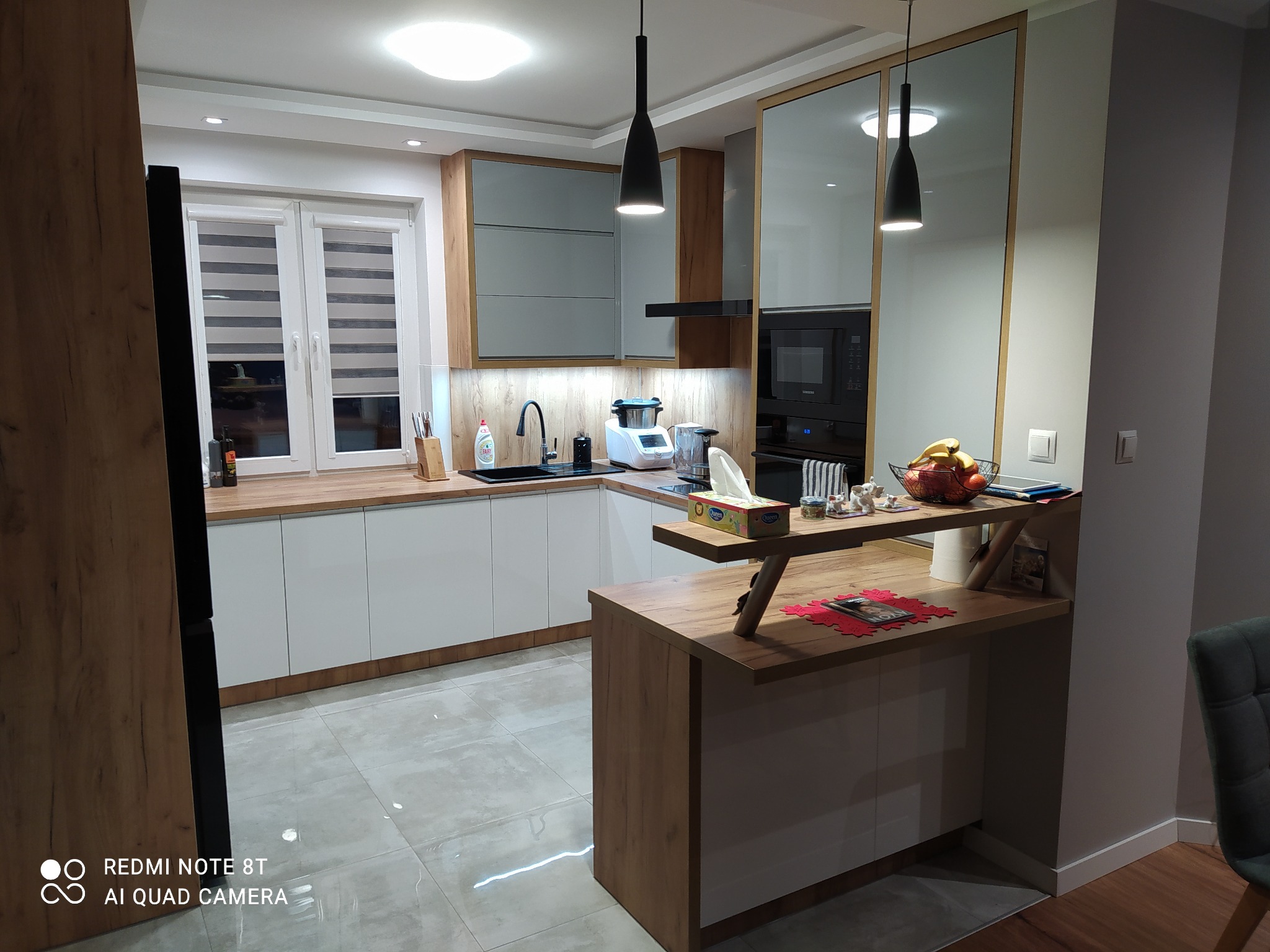 Nowoczesna kuchnia z białymi szafkami i drewnianymi blatami, oświetlona wiszącymi lampami, z wyspą kuchenną i elementami dekoracyjnymi.