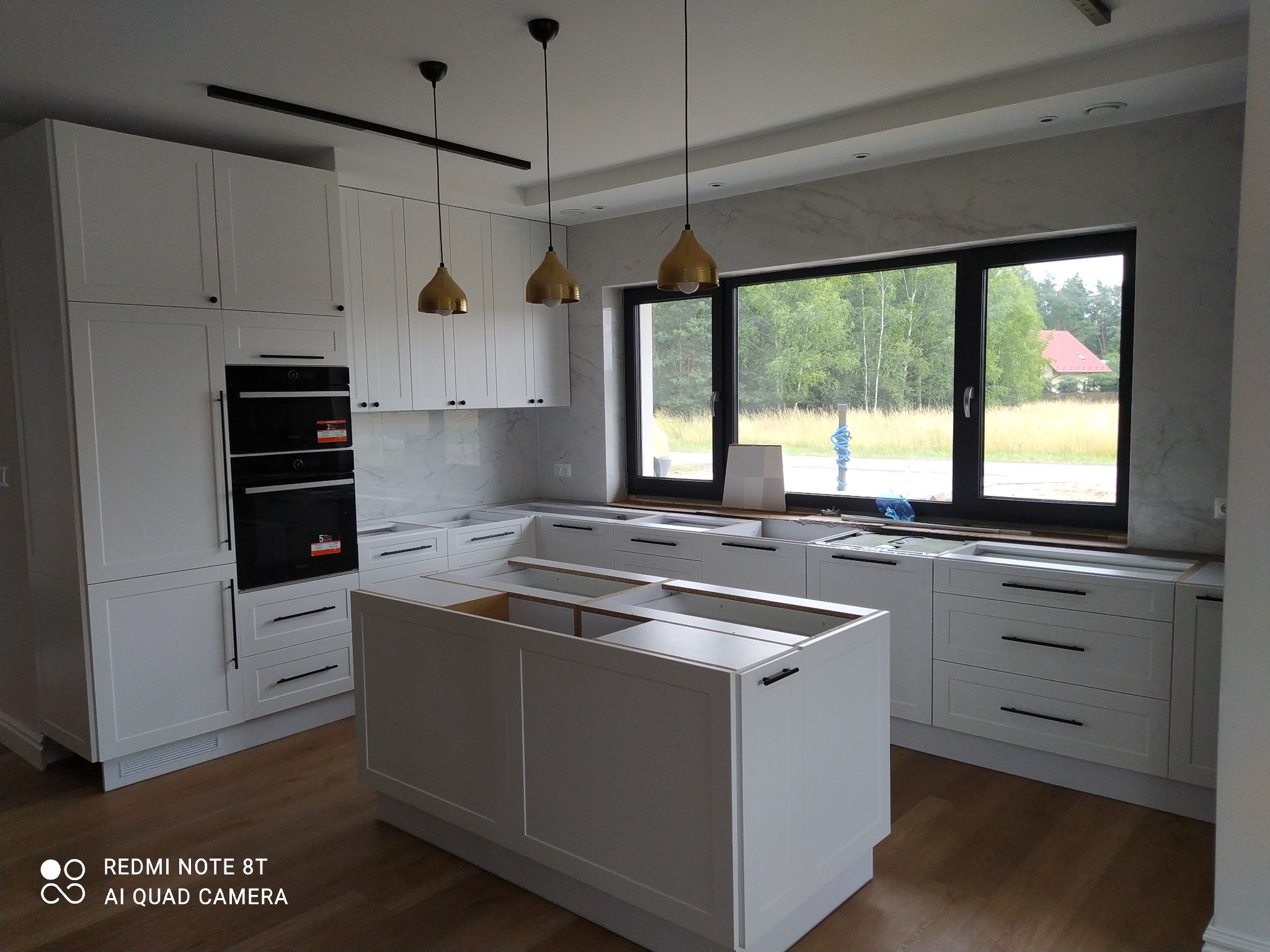 Biała kuchnia w trakcie montażu z wyspą kuchenną, widok na okno z widokiem na zieleń, złote lampy wiszące nad wyspą, marmurowe wykończenie ściany.