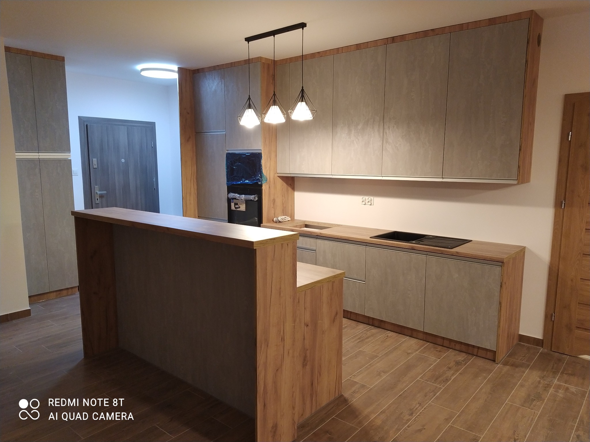 Nowoczesna kuchnia z szarymi frontami szafek i drewnianymi akcentami, oświetlona trzema wiszącymi lampami, z wyspą kuchenną i widocznym sprzętem AGD.