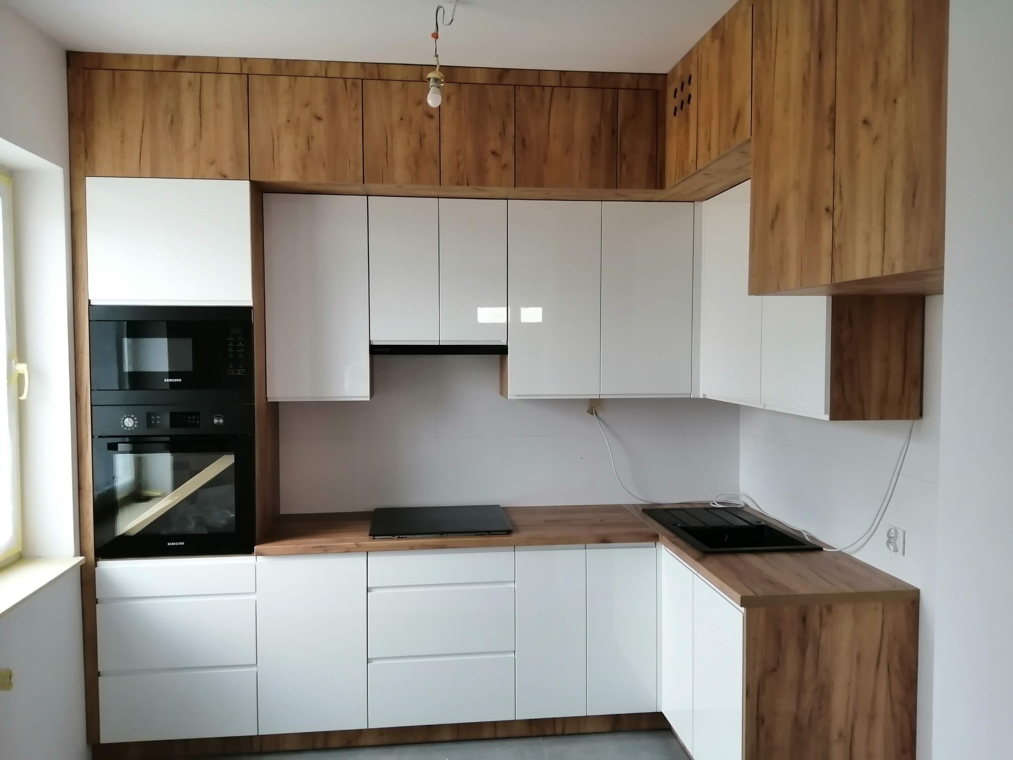 Nowoczesna kuchnia z białymi szafkami i drewnianymi blatami, płyta indukcyjna i piekarnik Samsung, surowa żarówka zwisająca z sufitu.
