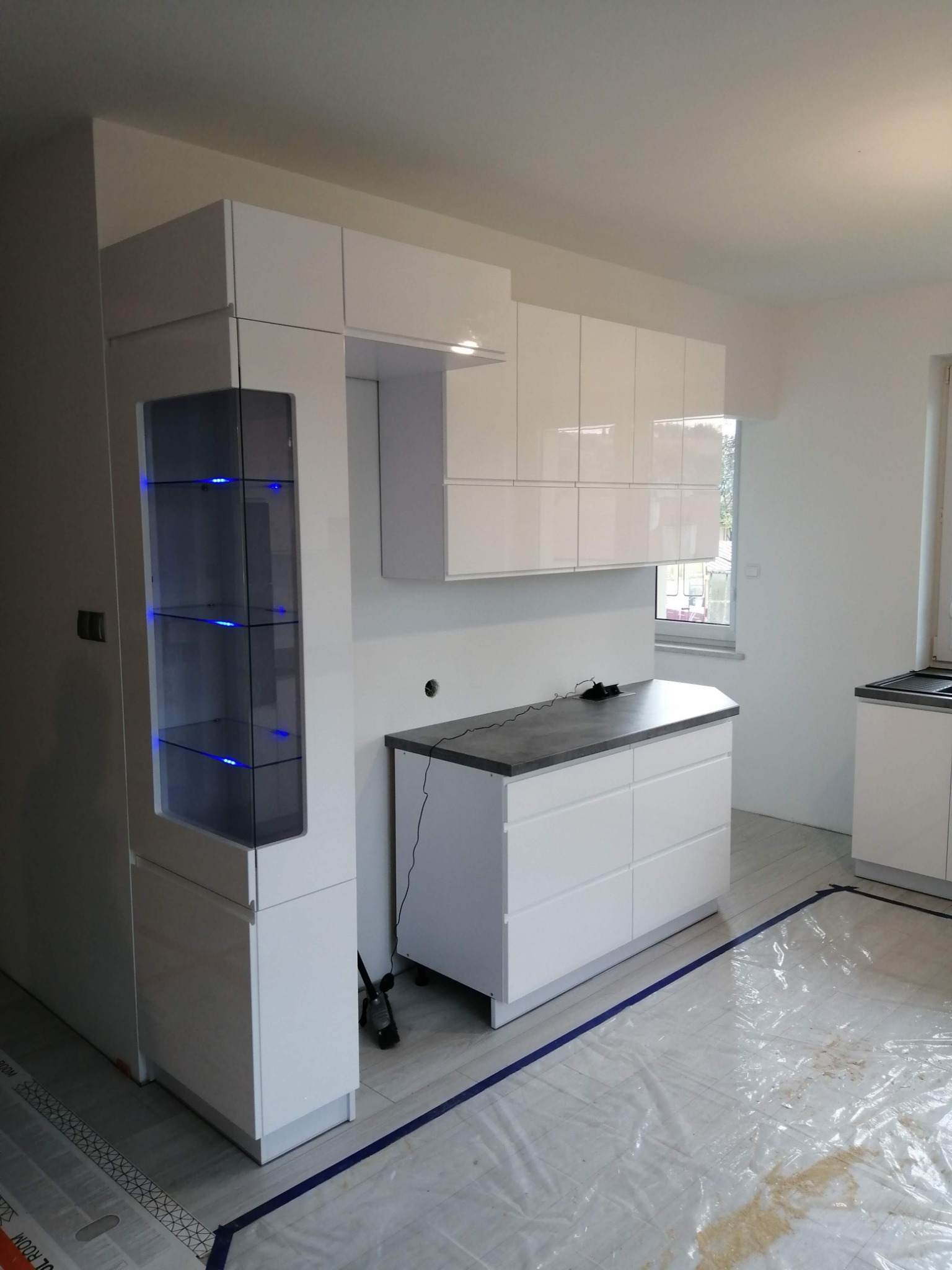 Nowoczesna, biała zabudowa kuchenna z podświetlaną witryną i szarym blatem, widoczne zabezpieczenia podłogi przed zabrudzeniem podczas montażu.