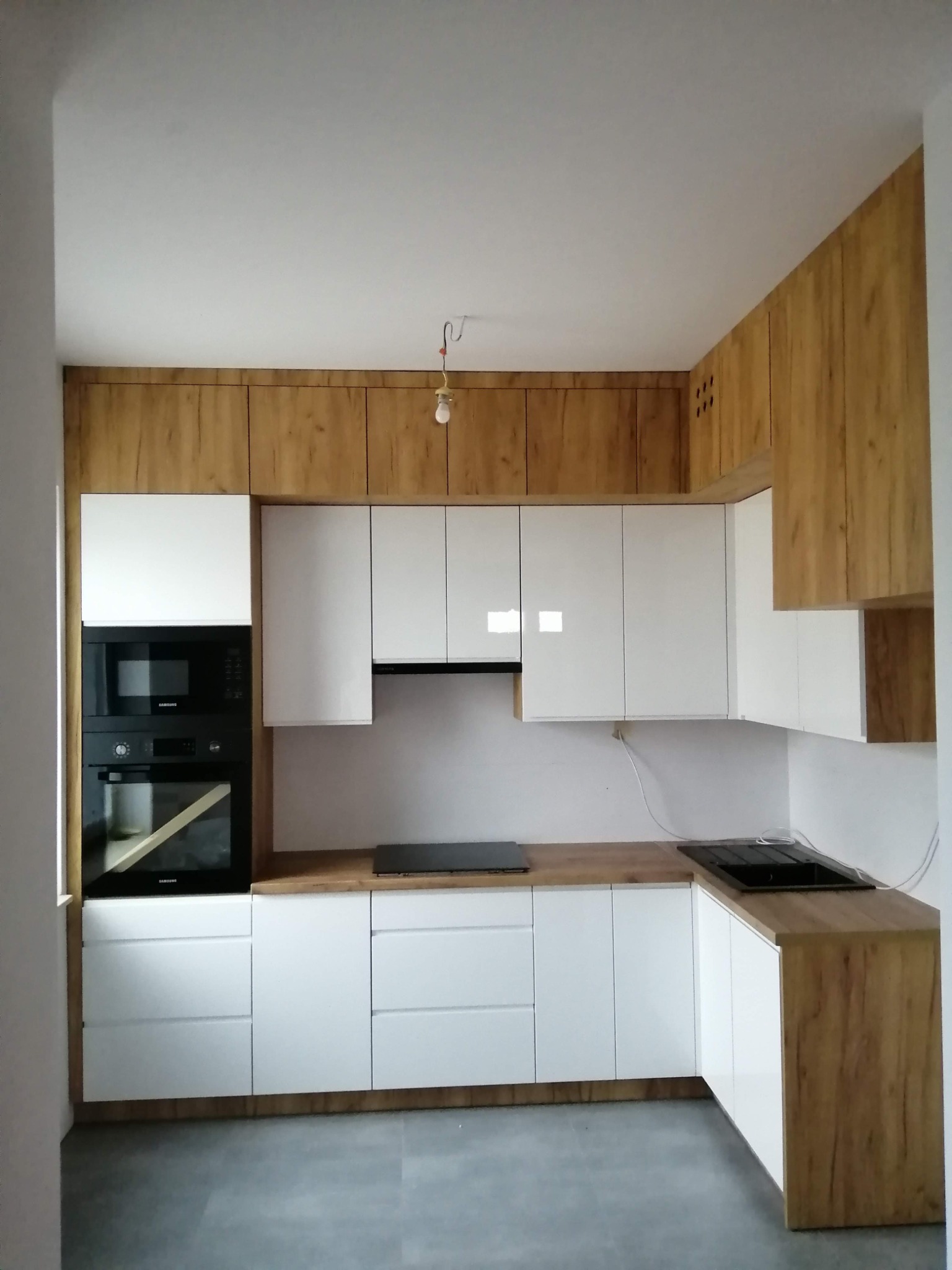 Nowoczesna, narożna zabudowa kuchenna z białymi, lakierowanymi frontami i drewnopodobnymi panelami górnymi i bocznymi, widoczny sprzęt AGD marki Samsung, surowy stan wykończenia wnętrza z wiszącą...
