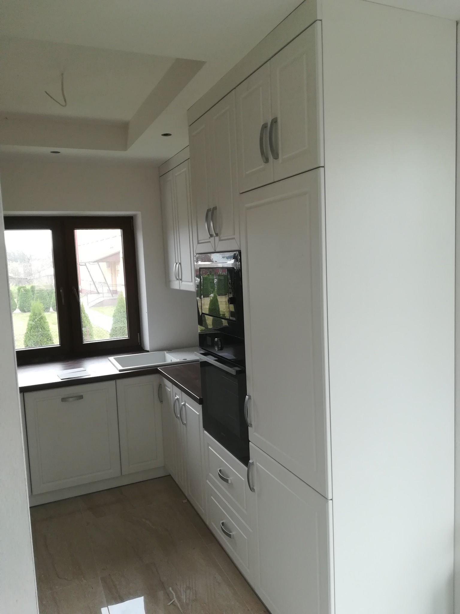 Jasna, nowoczesna zabudowa kuchenna z białymi frontami i ciemnym blatem, widok z boku, okno z widokiem na ogród w tle.