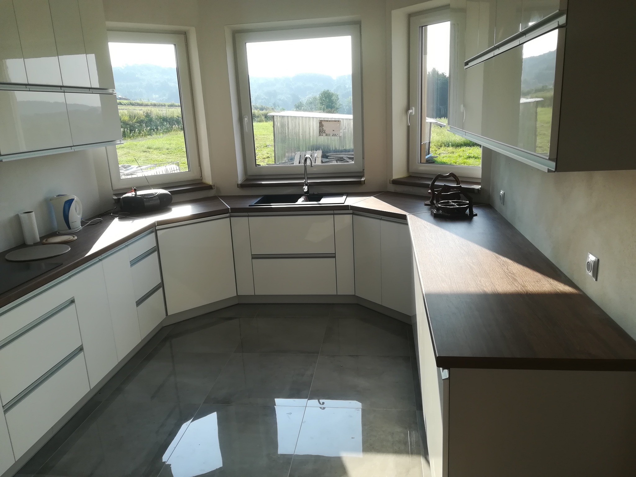 Jasna kuchnia na wymiar w kształcie litery U z drewnianym blatem, szarymi płytkami podłogowymi i widokiem na zieloną okolicę przez okna.