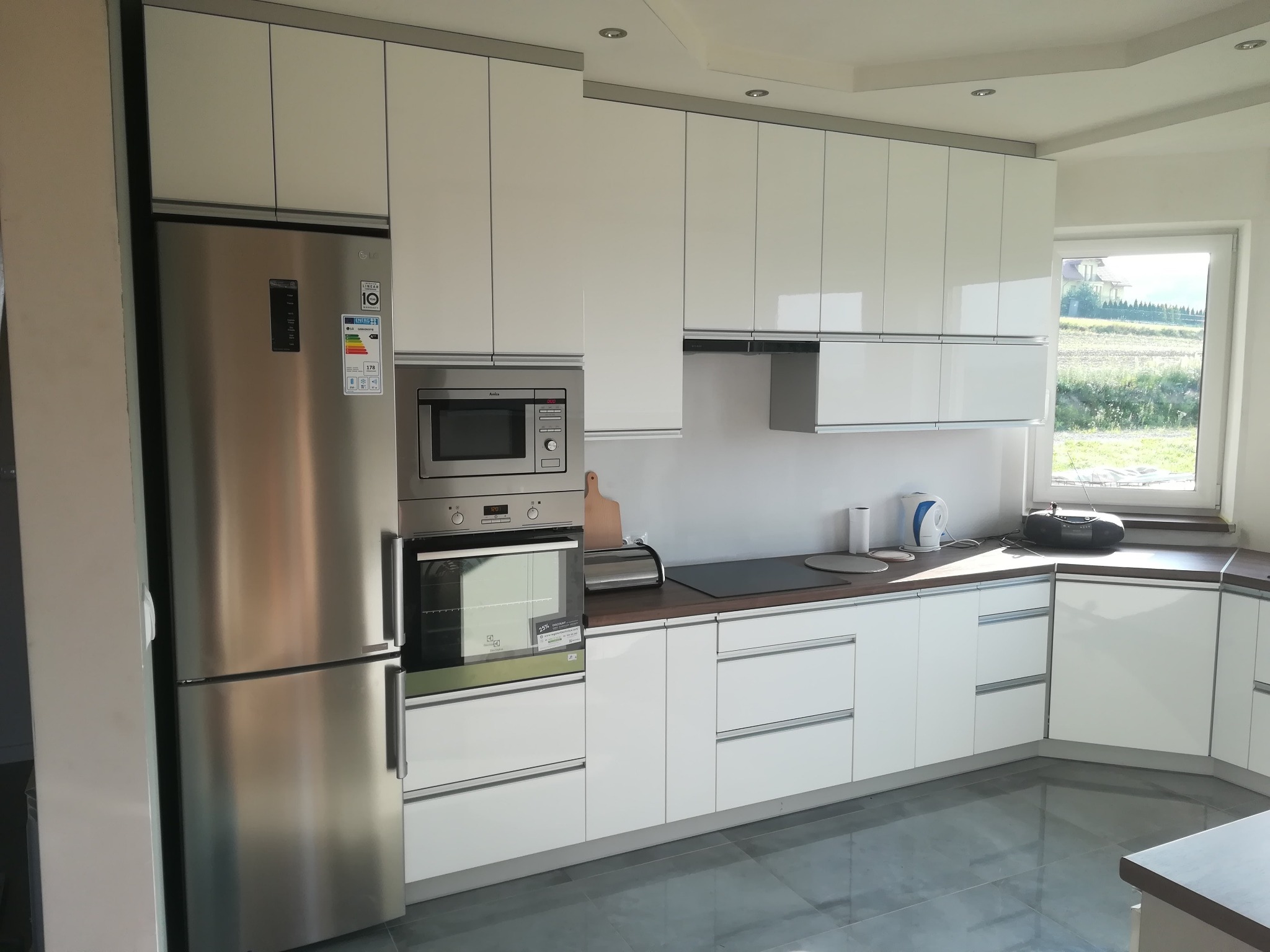 Jasna kuchnia z białymi, lakierowanymi frontami szafek, srebrnym wykończeniem, wbudowanym sprzętem AGD i szarymi płytkami podłogowymi, widok na ogród przez okno.