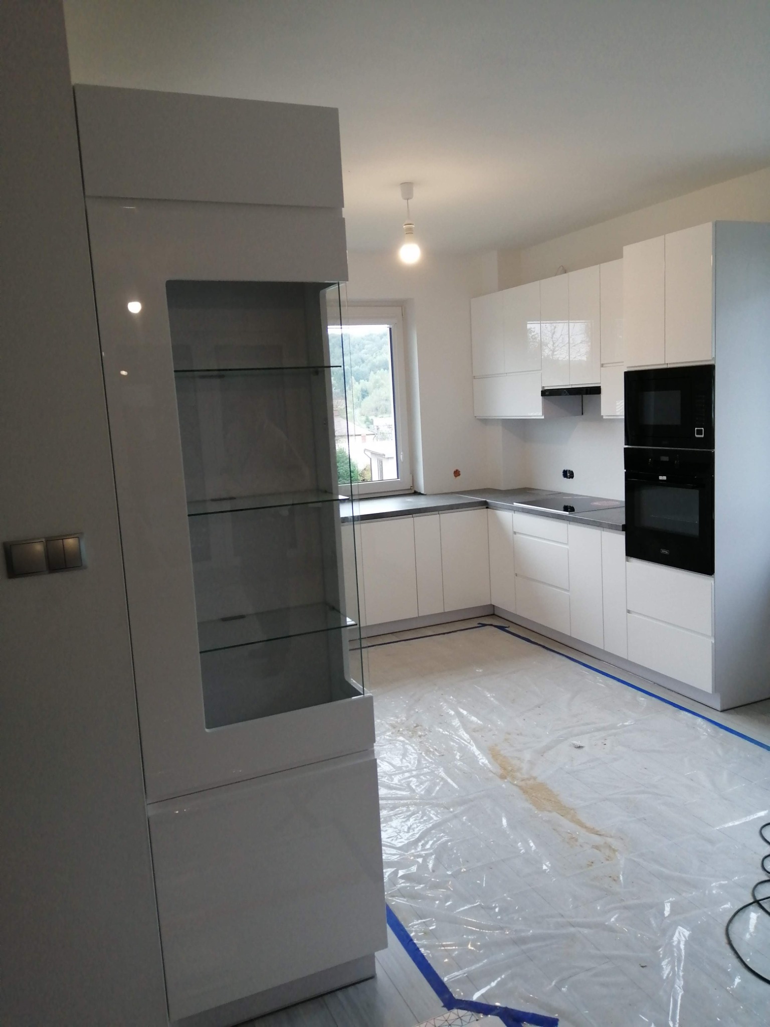 Nowoczesna, biała kuchnia z przeszklonym regałem, jeszcze w trakcie montażu, z folią ochronną na podłodze.