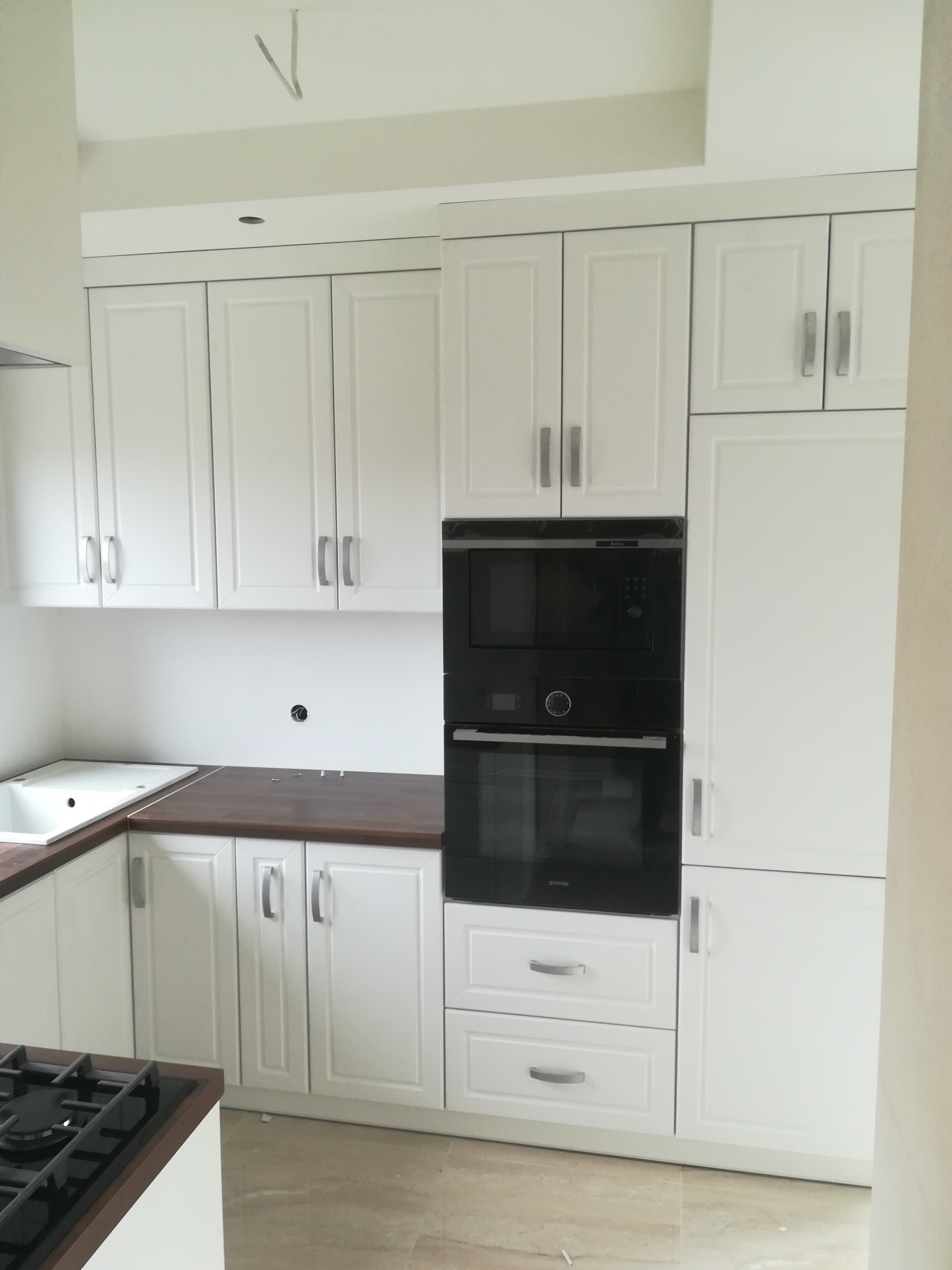 Biała zabudowa kuchenna z ciemnym blatem i wbudowanym piekarnikiem oraz mikrofalówką, widoczne niedokończone podłączenie elektryczne.