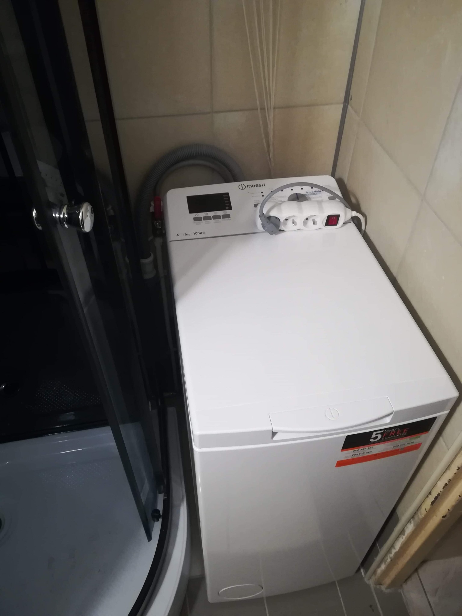 Wnętrze małej łazienki z kabiną prysznicową i pralką Indesit z podłączonym przedłużaczem, widoczne płytki ceramiczne na ścianach.