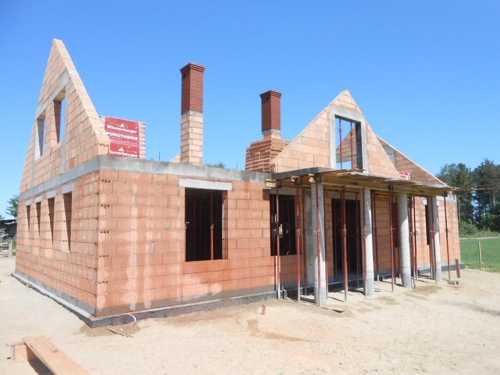 Wznoszenie ścian parterowego domu z cegły Porotherm, widoczne kominy z cegły klinkierowej i podpory stropu nad wejściem, w tle błękitne niebo i zieleń drzew.