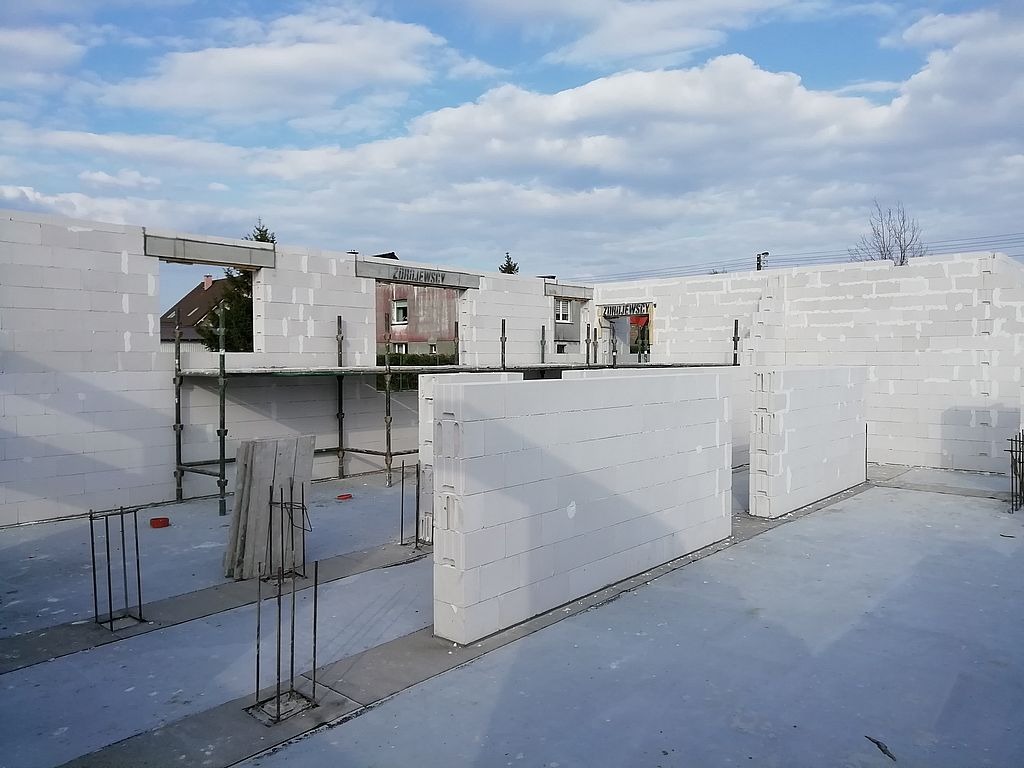 Wznoszenie ścian domu jednorodzinnego z białych bloczków, widoczne zbrojenia i podpory, w tle budynek z napisem 'ZURAJEWSCY'.