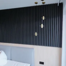 Nowoczesna sypialnia w Strzelcach Wielkich z czarną ścianą z pionowych paneli i minimalistycznymi złotymi lampami wiszącymi nad łóżkiem z szarym zagłówkiem.