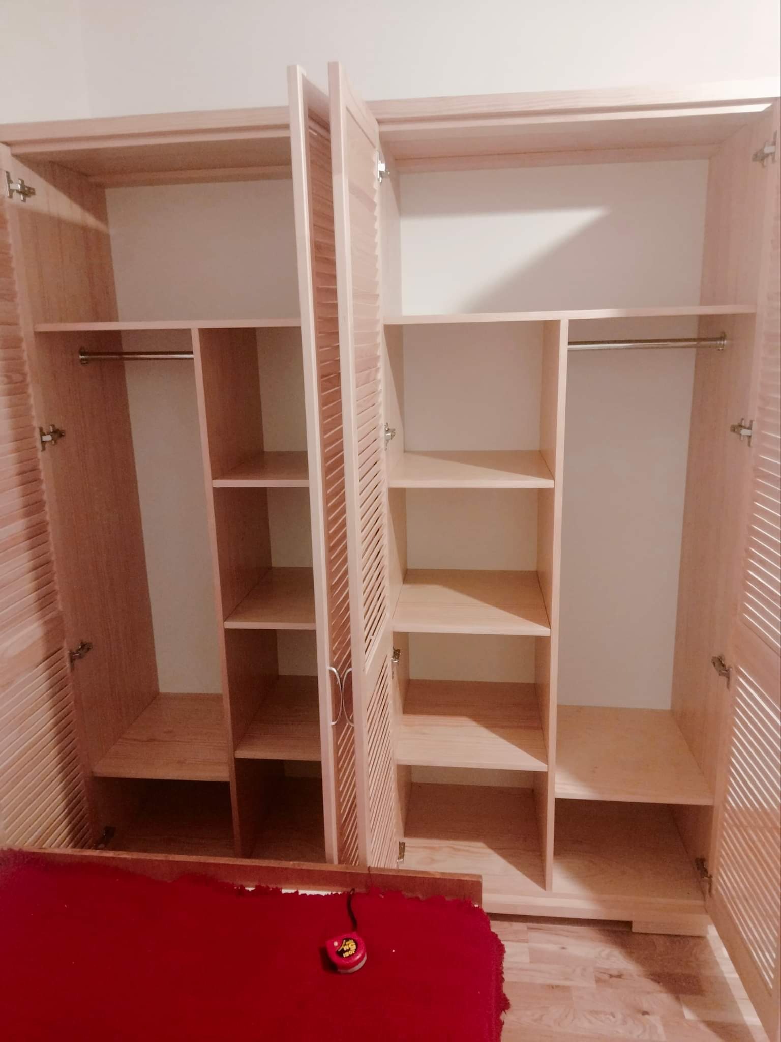 Wnętrze jasnej, drewnianej szafy z lamelkowymi drzwiami, widoczne półki i drążek na ubrania, z boku leży zwinięta miarka.