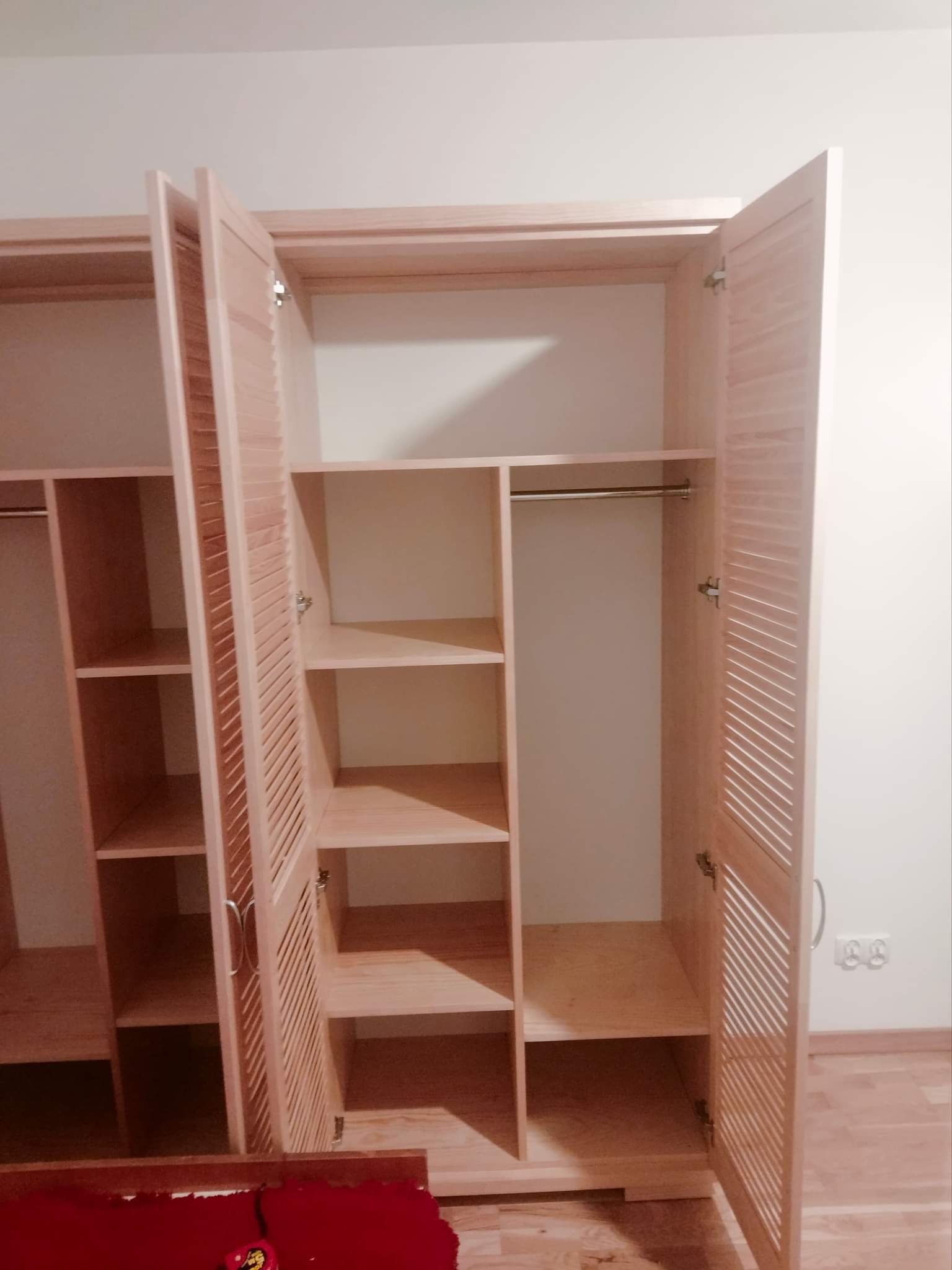 Jasna drewniana szafa z lamelowanymi drzwiami, otwarte skrzydła ukazują puste półki i drążek na ubrania, widok na wnętrze szafy.