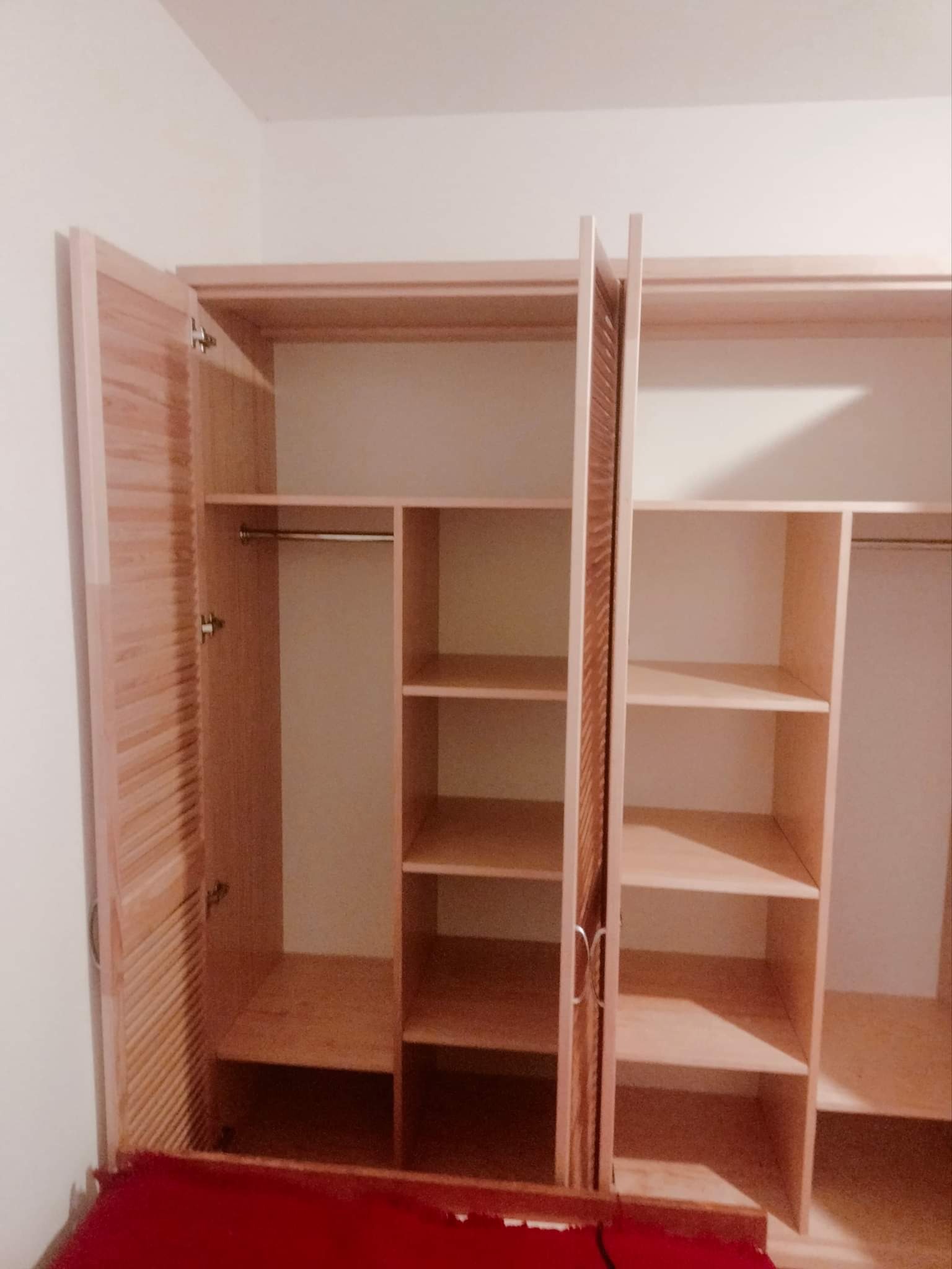 Jasna drewniana szafa trzydrzwiowa z otwartymi lamelowymi drzwiami po lewej stronie, widoczne półki i drążek na ubrania.