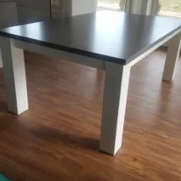 Stół z ciemnym blatem i białymi nogami na drewnianej podłodze, widoczny fragment okna w tle.