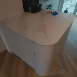 Nowoczesna wyspa kuchenna z białym blatem imitującym marmur i zaokrąglonymi bokami, na blacie leżą narzędzia i skrzynki narzędziowe, podłoga wyłożona parkietem ułożonym w jodełkę.