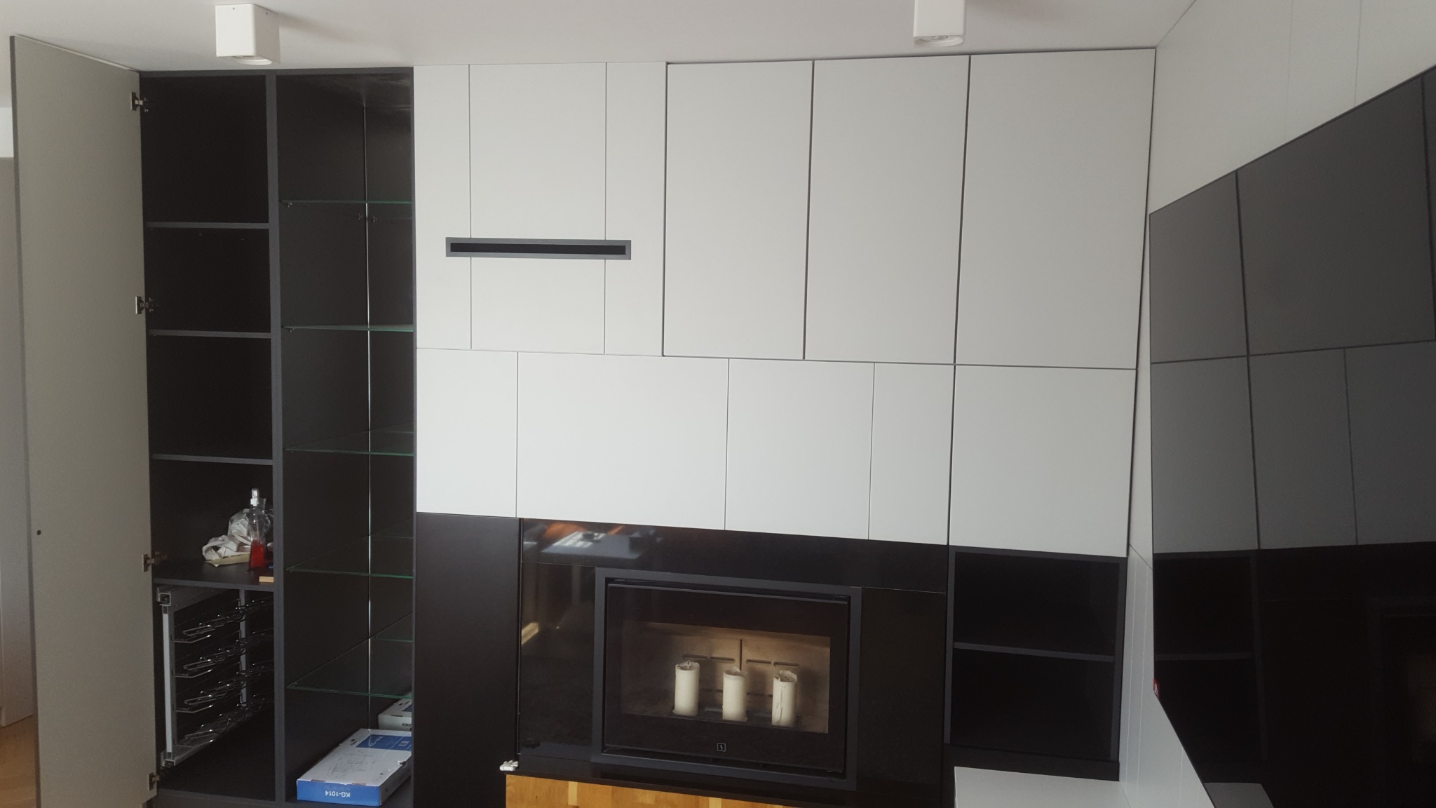 Nowoczesna zabudowa ściany z szafkami w kolorach bieli i grafitu, z otwartą szafką z szklanymi półkami po lewej stronie i kominkiem elektrycznym z trzema świecami w centralnej części.