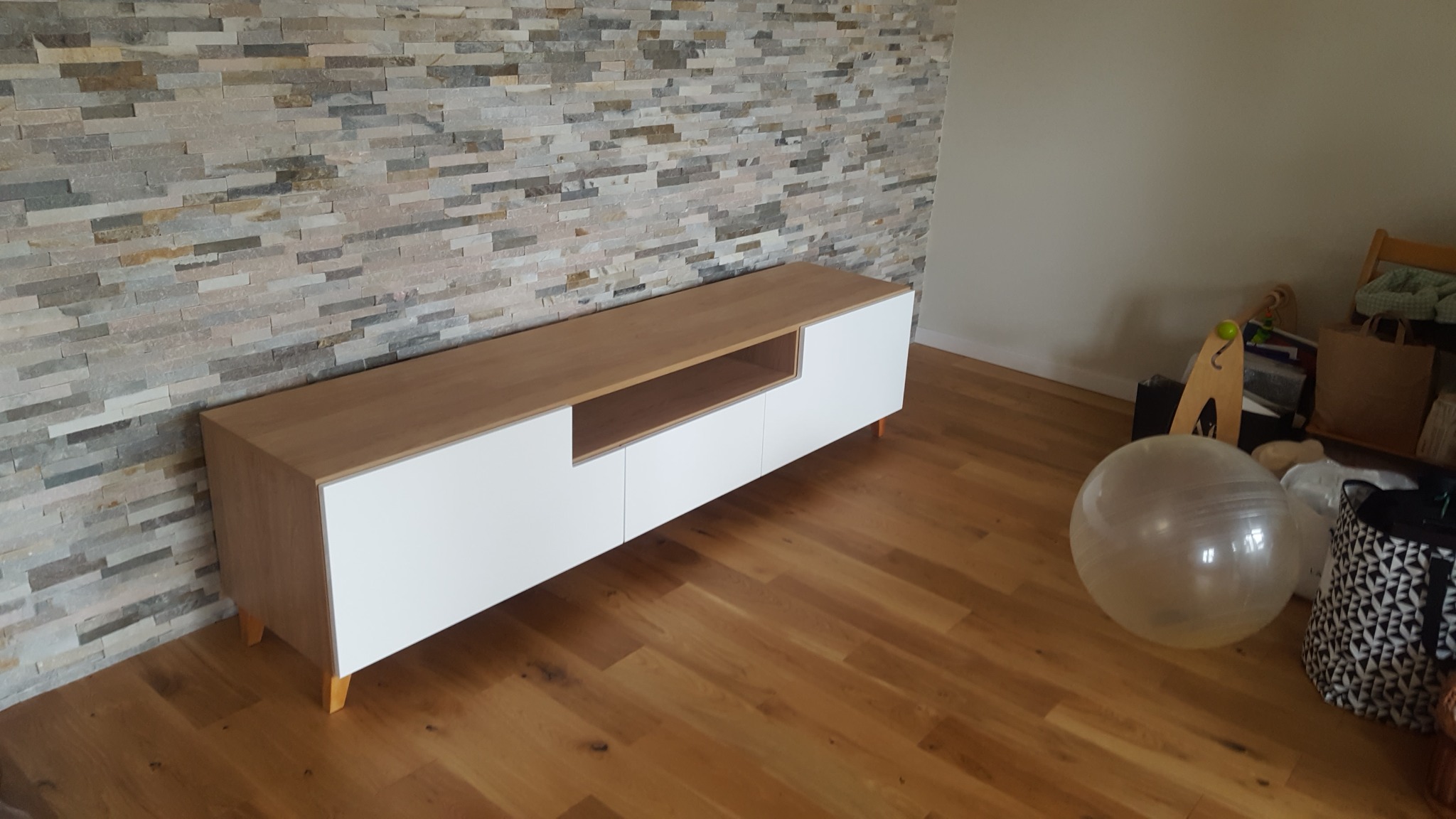 Długa szafka RTV z białymi frontami i drewnianym blatem, umieszczona na drewnianej podłodze przy ścianie wyłożonej dekoracyjnym kamieniem.