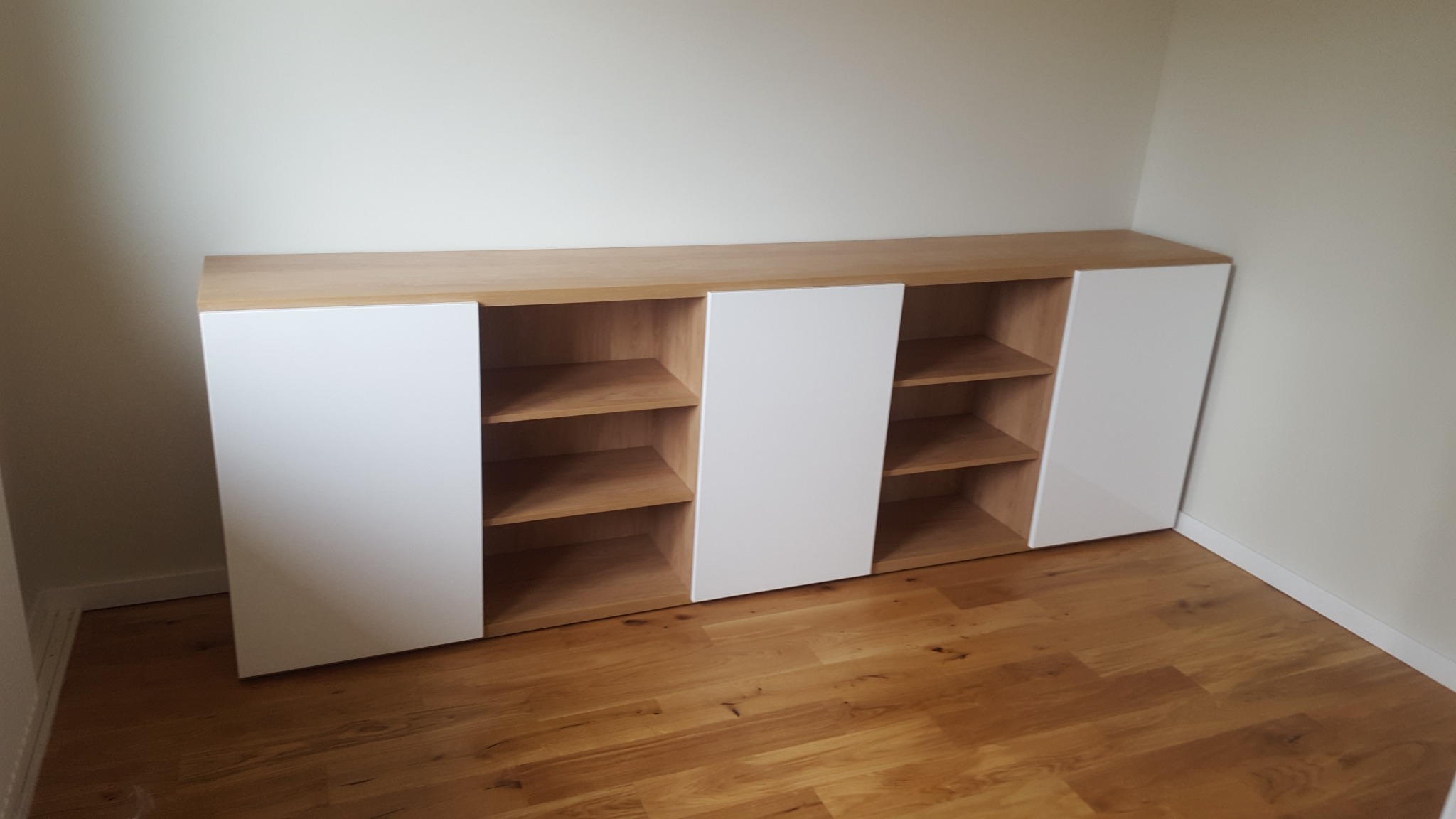 Niska szafka z białymi frontami i drewnianym blatem, z otwartymi półkami, ustawiona na drewnianej podłodze w rogu pokoju.