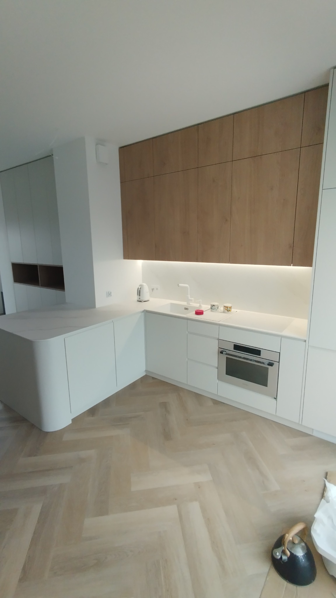 Nowoczesna kuchnia z białymi frontami i drewnianymi szafkami wiszącymi, oświetlenie LED pod szafkami, wyspa kuchenna z zaokrąglonym blatem, parkiet w jodełkę.