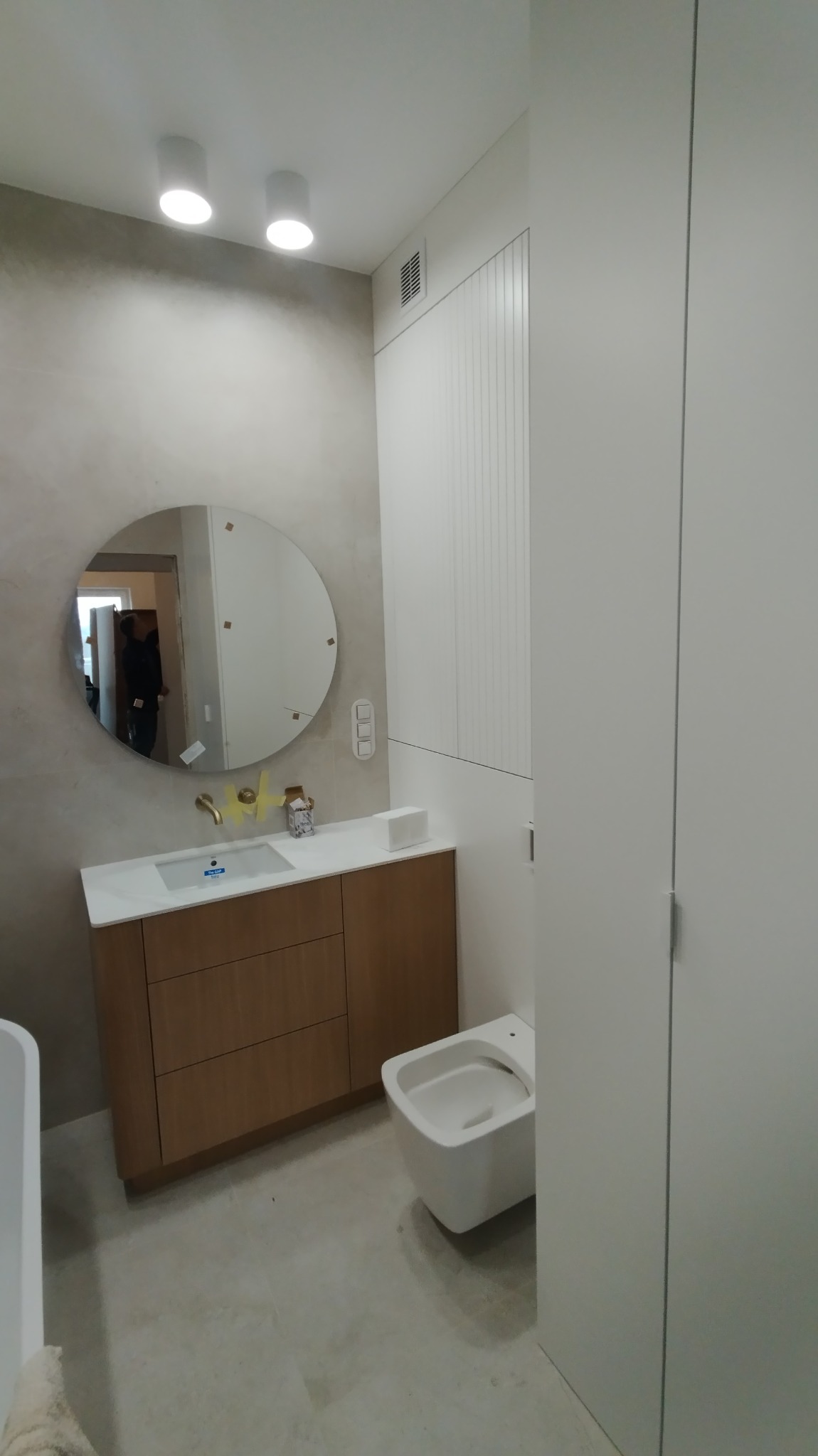 Nowoczesna łazienka z okrągłym lustrem, umywalką nablatową i szafką z drewnopodobnym frontem. Białe ściany i toaleta podwieszana. Minimalistyczne oświetlenie sufitowe.