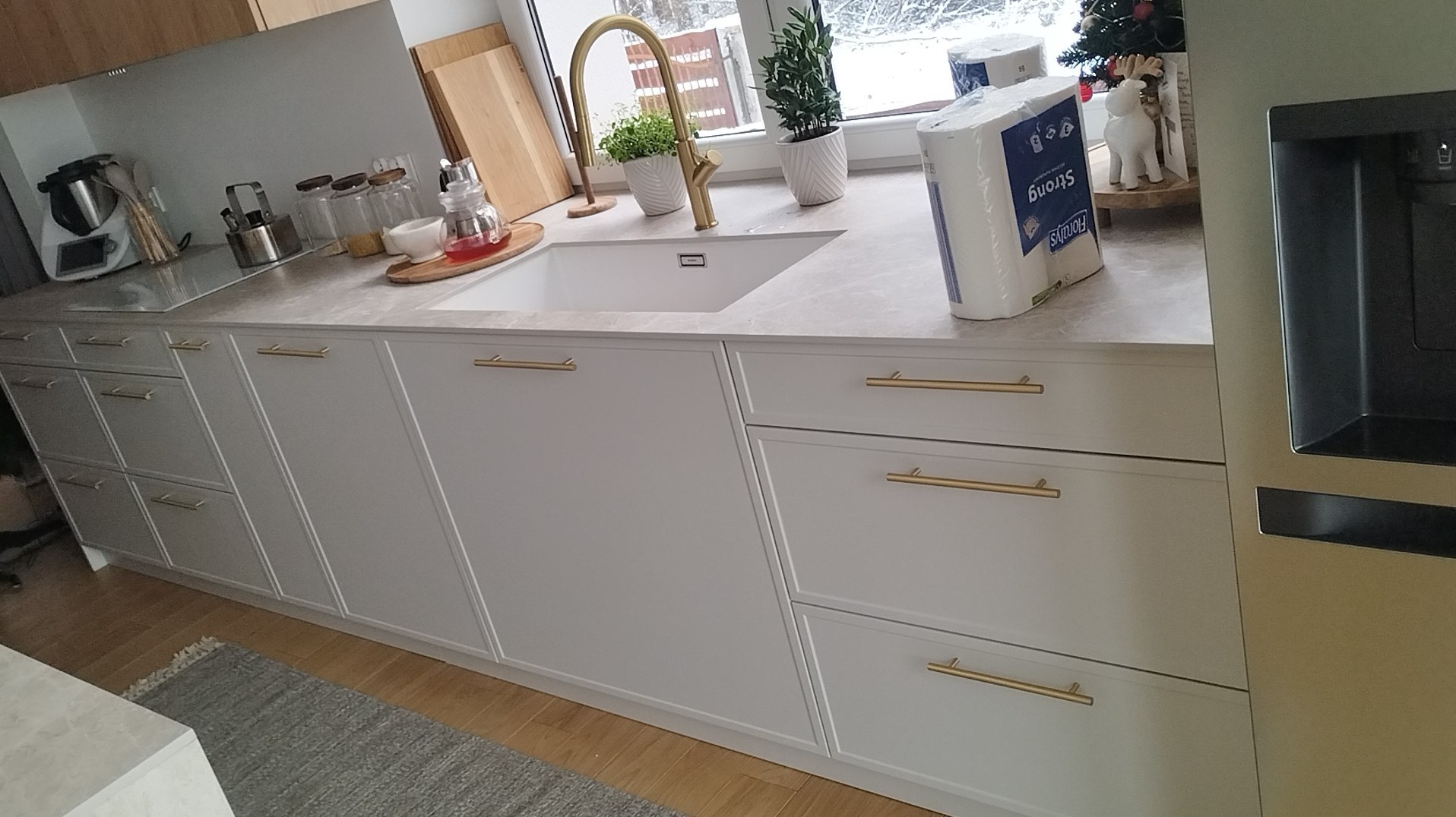 Jasna kuchnia z białymi frontami szafek z frezowanymi ramkami i złotymi uchwytami, marmurowy blat, złota bateria przy zlewie, akcesoria kuchenne i dekoracje na blacie.