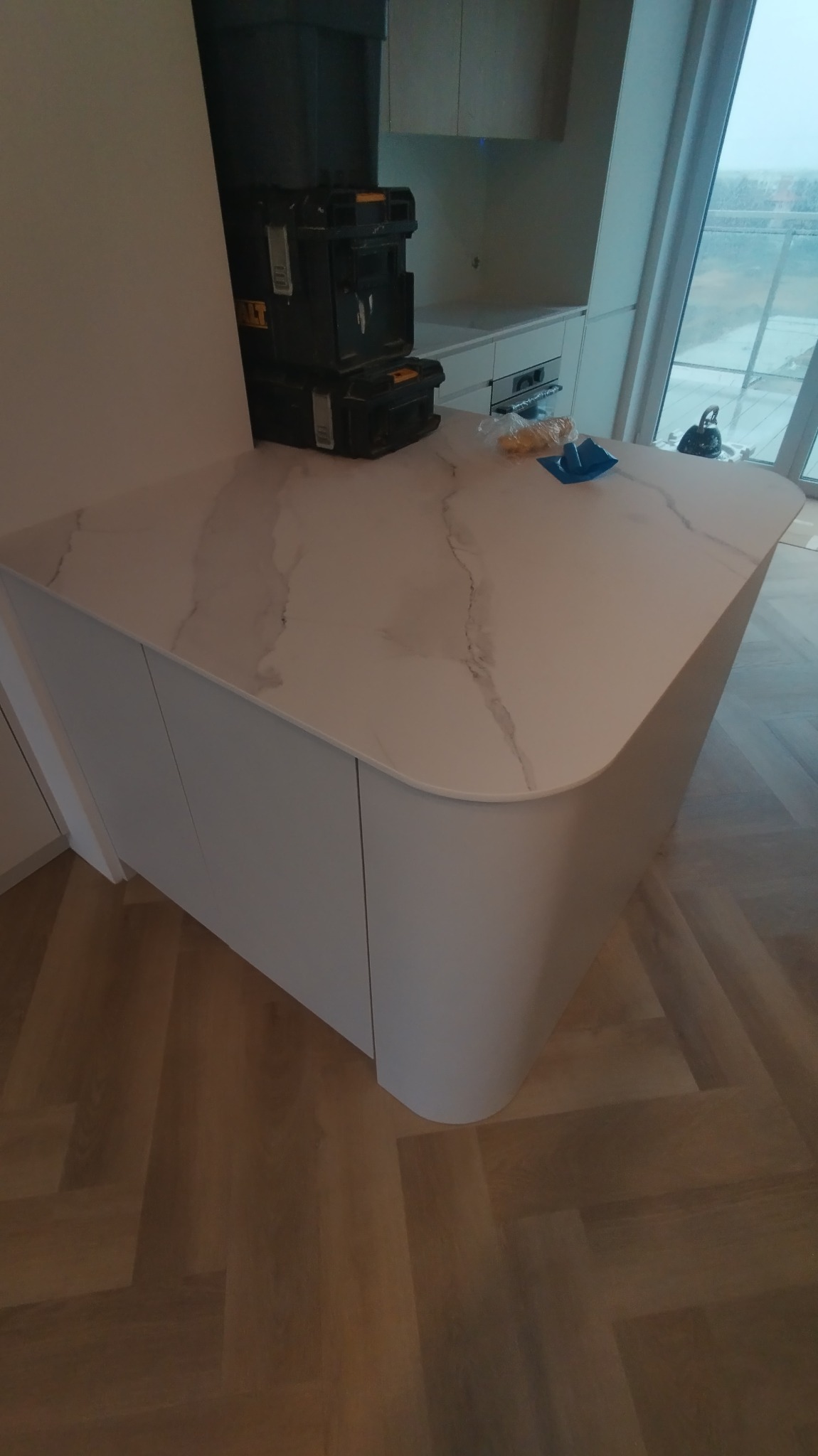 Nowoczesna wyspa kuchenna z białym blatem imitującym marmur i zaokrąglonymi bokami, na blacie leżą narzędzia i skrzynki narzędziowe, podłoga wyłożona parkietem ułożonym w jodełkę.