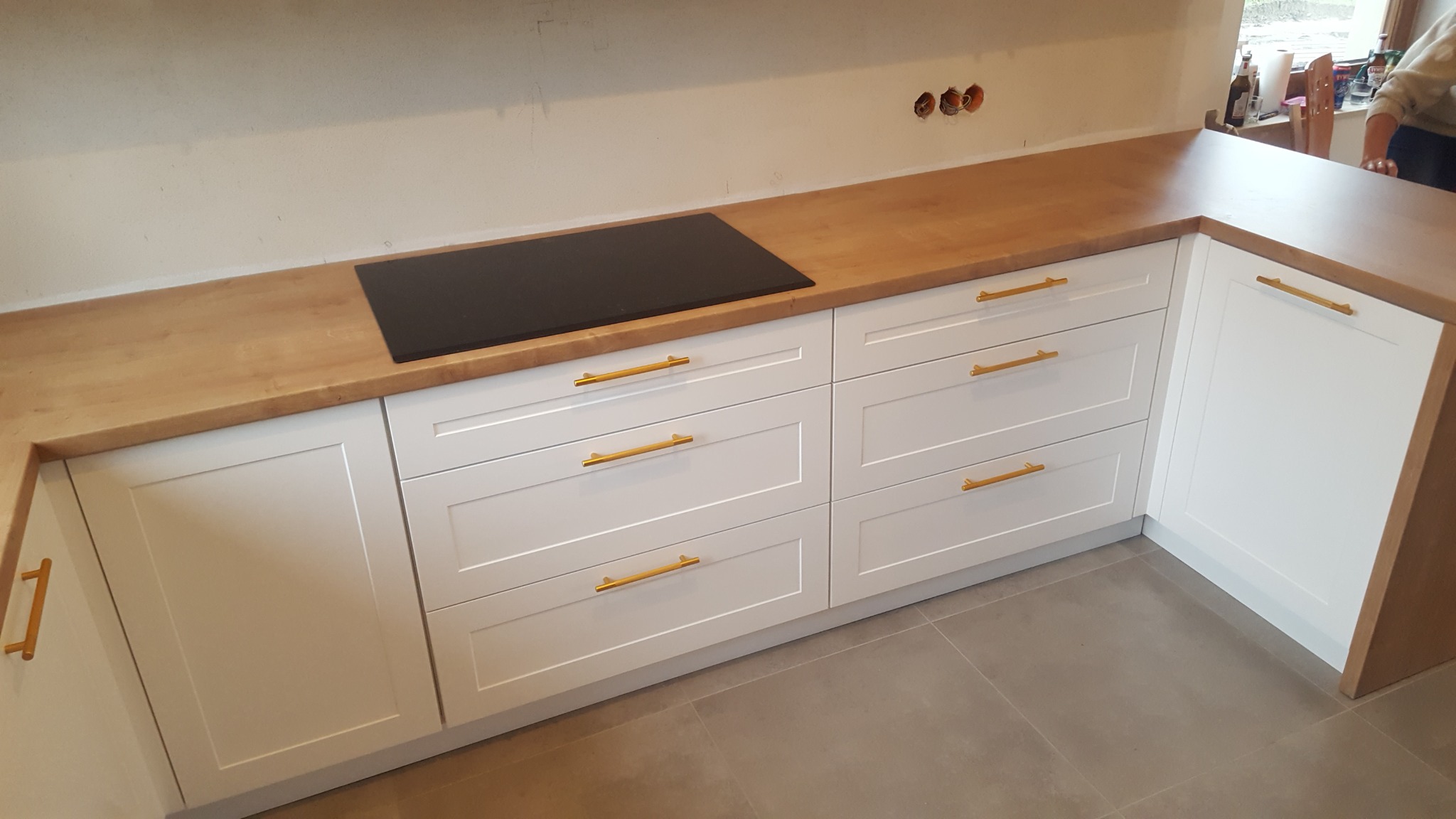 Białe szafki kuchenne z drewnianym blatem i złotymi uchwytami, z czarną płytą indukcyjną wbudowaną w blat, widoczne niedokończone gniazdka elektryczne w ścianie.