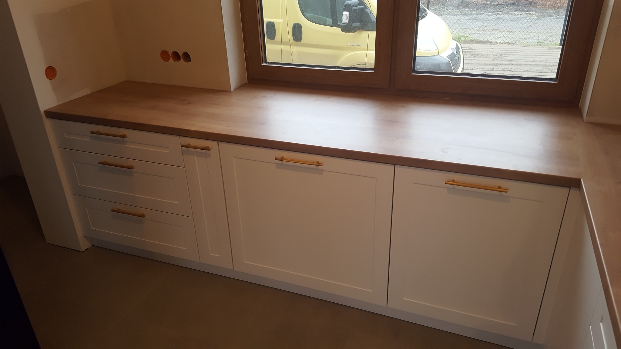 Białe szafki kuchenne z drewnianym blatem pod oknem, widoczny żółty samochód za oknem, minimalistyczne złote uchwyty.