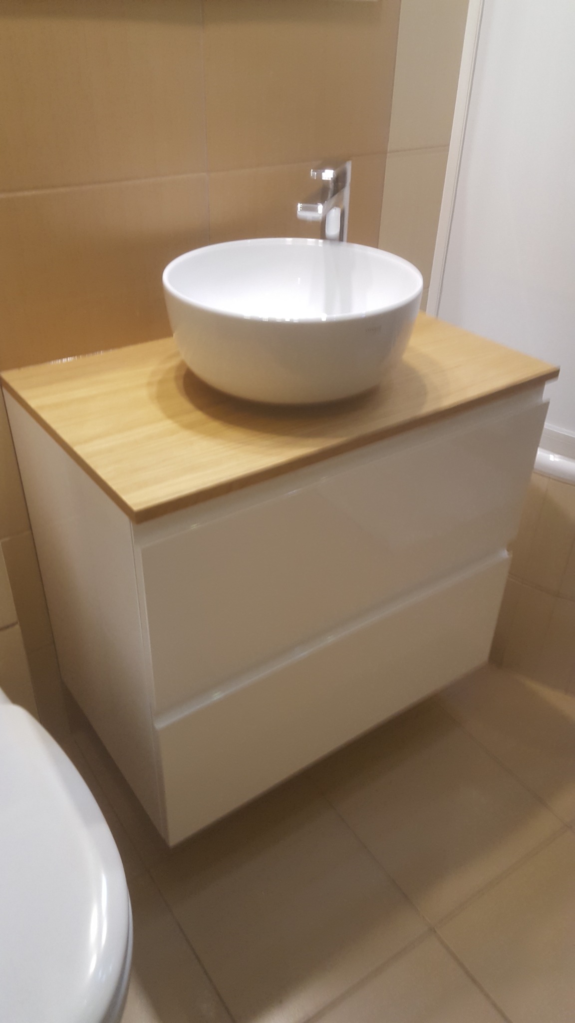 Biała, okrągła umywalka nablatowa z chromowaną baterią na szafce z dwoma szufladami i blatem z jasnego drewna, widok z boku, jasne wnętrze łazienki.