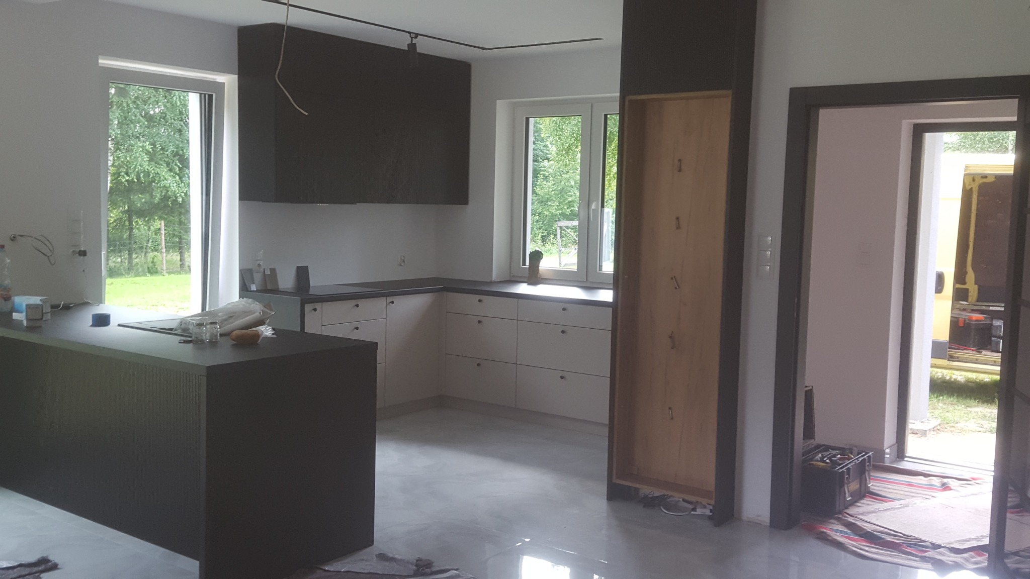 Wnętrze kuchni w trakcie montażu: widoczne niedokończone szafki, wyspa kuchenna z ciemnym blatem, okno z widokiem na zieleń i narzędzia w przejściu do innego pomieszczenia.