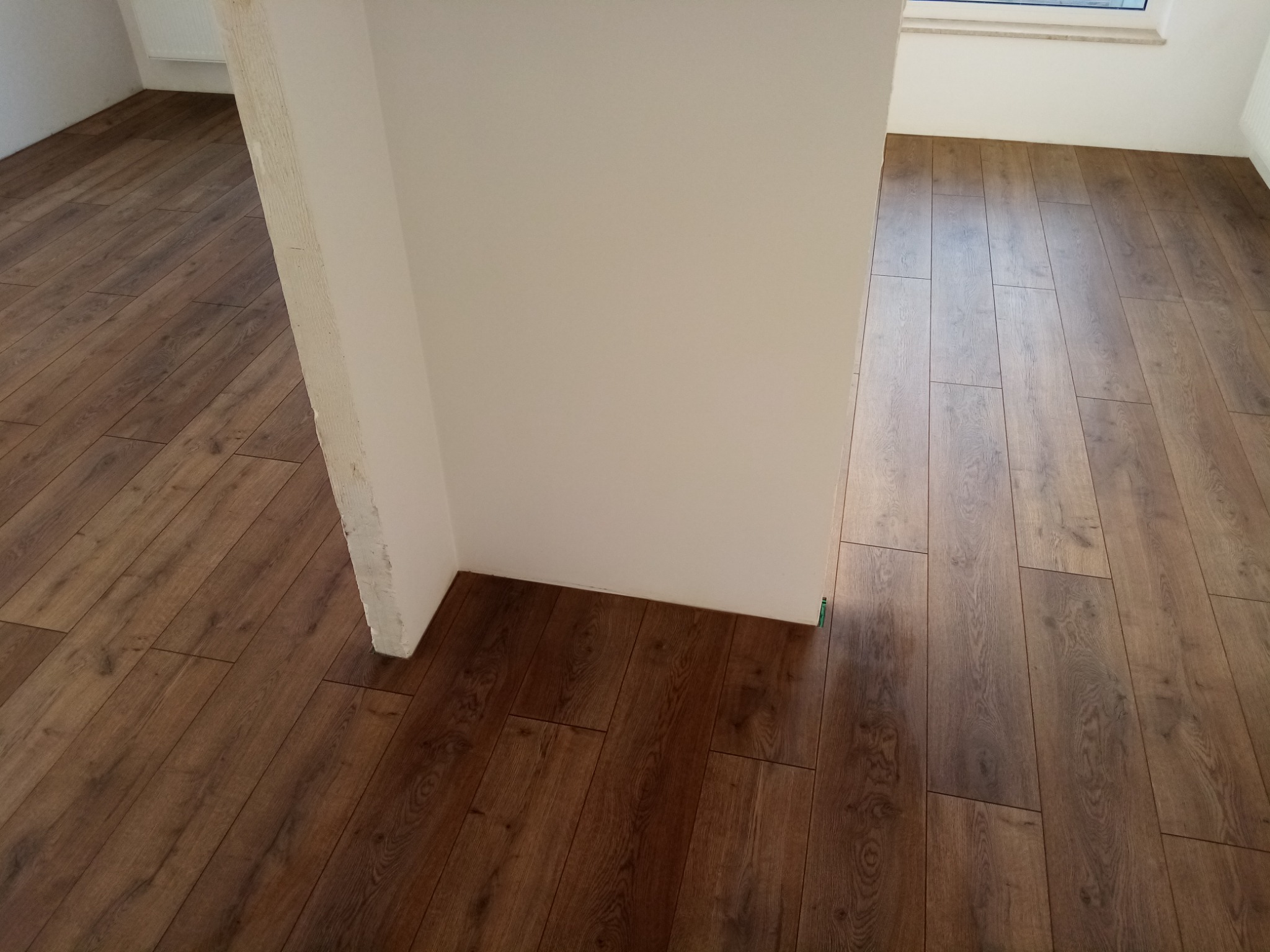 Ułożona podłoga z paneli o ciemnym, drewnopodobnym wzorze w nowym pomieszczeniu, widoczny fragment ściany i okno w tle.