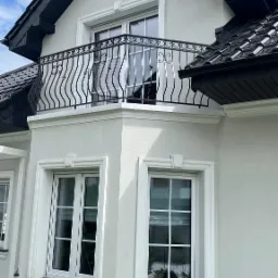 Biały dom z czarnym dachem i balkonem z czarną, ozdobną balustradą, widok z dołu na narożnik budynku.