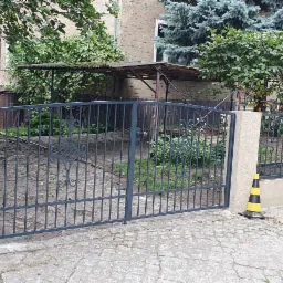 Nowoczesna, szara brama wjazdowa z metalowymi prętami i ozdobnym elementem, ustawiona między betonowymi słupkami, zabezpieczona dwoma żółto-czarnymi pachołkami ostrzegawczymi na brukowanym podjeździe.