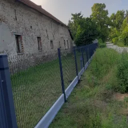 Nowoczesne ogrodzenie panelowe z podmurówką betonową, przylegające do starego, ceglanego budynku gospodarczego w wiejskim otoczeniu.