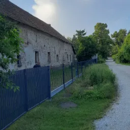 Szary, drewniany płot panelowy i metalowa siatka ogrodzeniowa wzdłuż drogi żwirowej obok starego, ceglanego budynku gospodarczego z widocznymi ubytkami tynku.