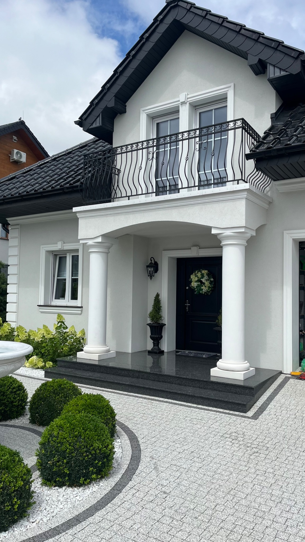 Elegancki dom we Wrocławiu z czarną, kutą balustradą balkonową, kolumnami przy wejściu i dekoracyjnym brukiem na podjeździe.