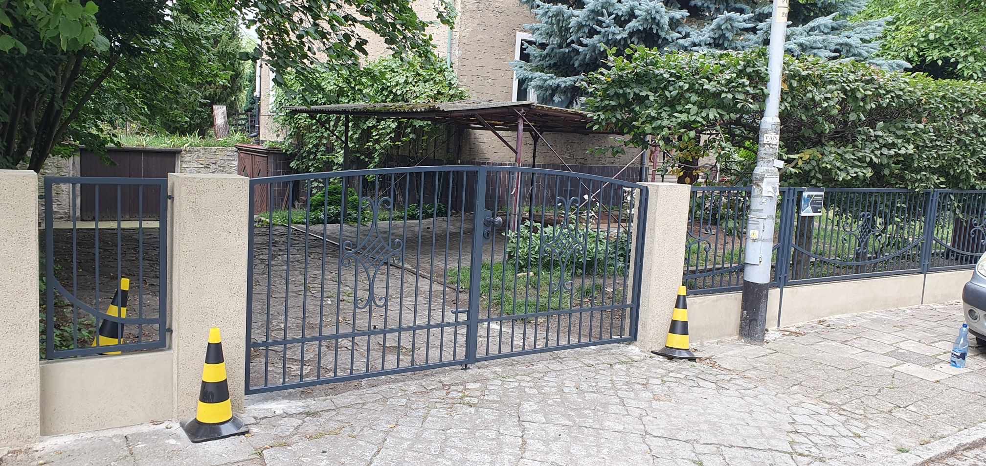 Nowoczesna, szara brama wjazdowa z metalowymi prętami i ozdobnym elementem, ustawiona między betonowymi słupkami, zabezpieczona dwoma żółto-czarnymi pachołkami ostrzegawczymi na brukowanym podjeździe.