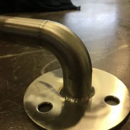 Metalowa rura wygięta pod kątem prostym, przyspawana do okrągłej podstawy z dwoma otworami, widoczne ślady spawania.