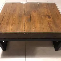 Kwadratowy stolik kawowy z blatem z dwóch grubych desek o wyraźnym usłojeniu, zabezpieczony szkłem, na czarnych, metalowych nogach w kształcie litery V.