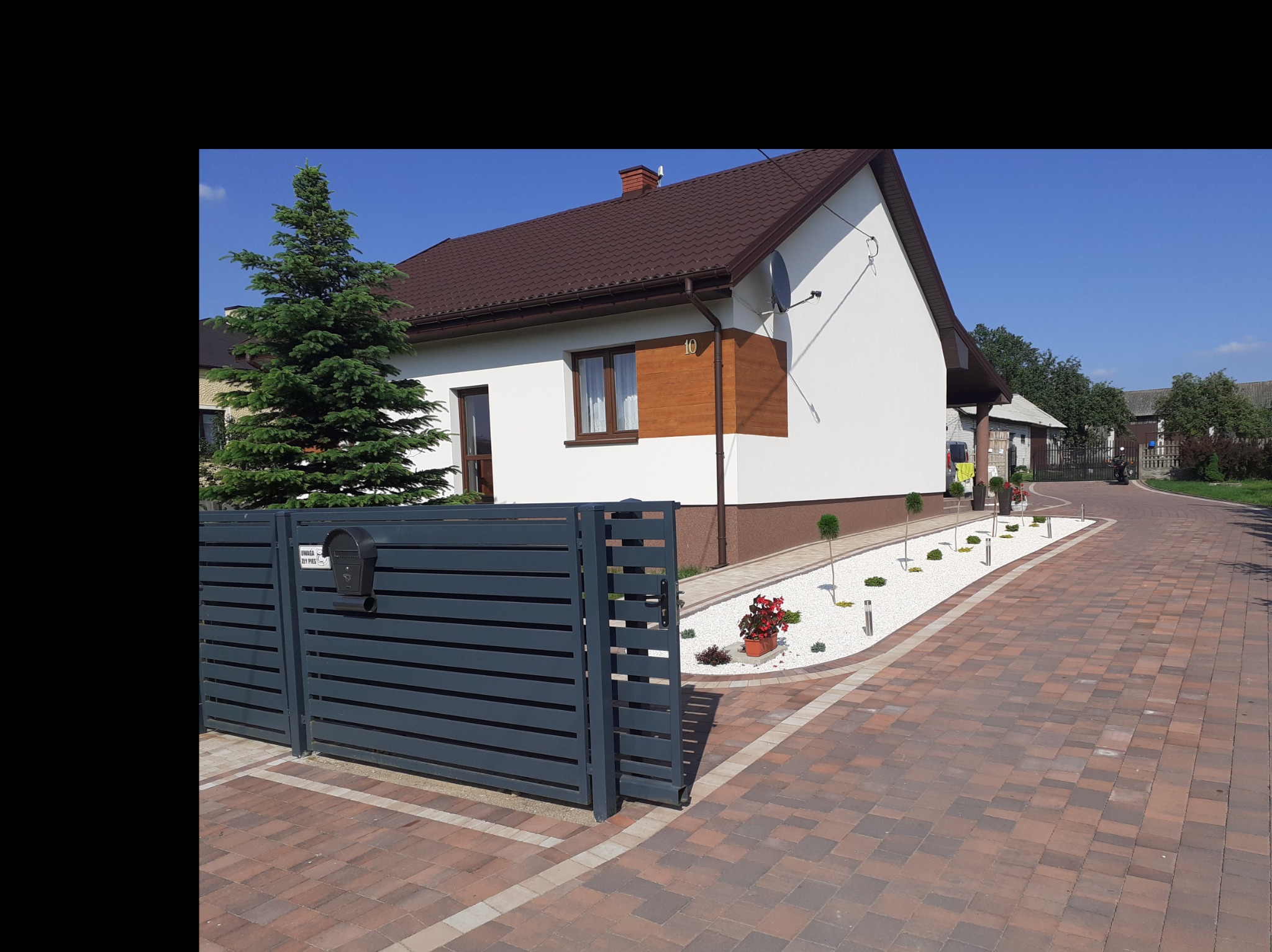 Nowoczesny dom jednorodzinny z białą elewacją, brązowym dachem i drewnianymi akcentami, widoczny zza szarej, poziomej bramy, z zadbanym ogrodem z białymi kamieniami i niskimi krzewami.