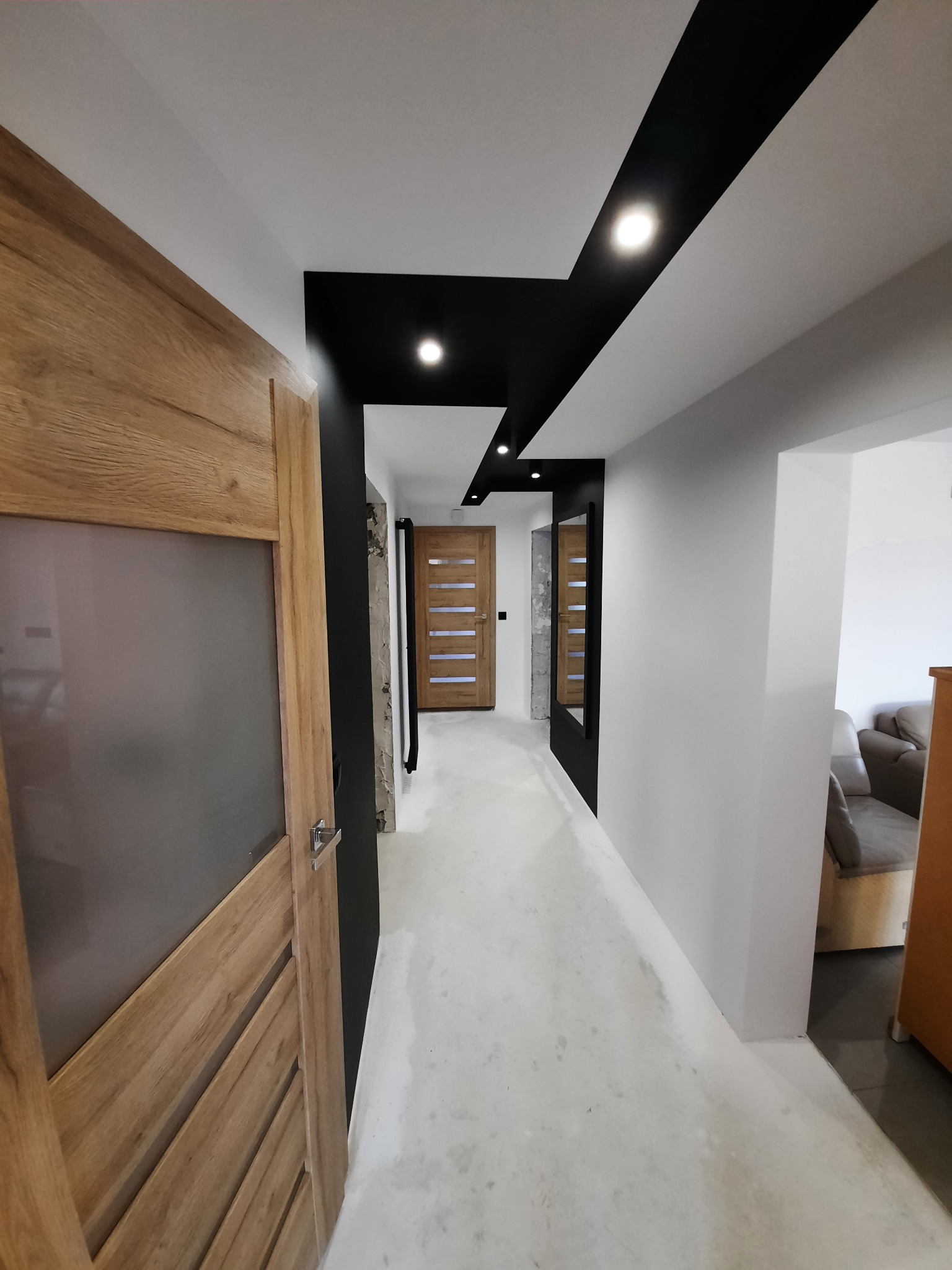 Korytarz w trakcie wykańczania: widoczne drzwi z szybą, czarne pasy na suficie z oświetleniem punktowym, surowa podłoga przed położeniem parkietu.