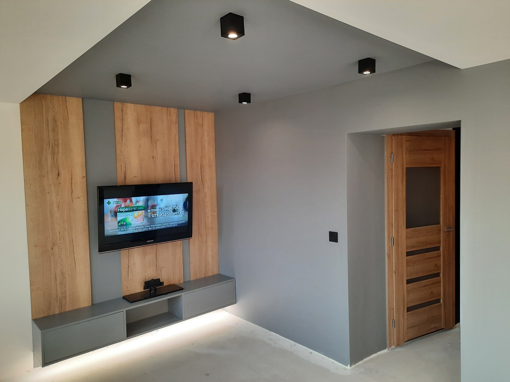 Nowoczesny salon w szarościach z akcentami drewna: ściana TV z podświetleniem LED, drewniane panele dekoracyjne, wisząca szafka RTV, drewniane drzwi z przeszkleniami i punktowe oświetlenie sufitowe.
