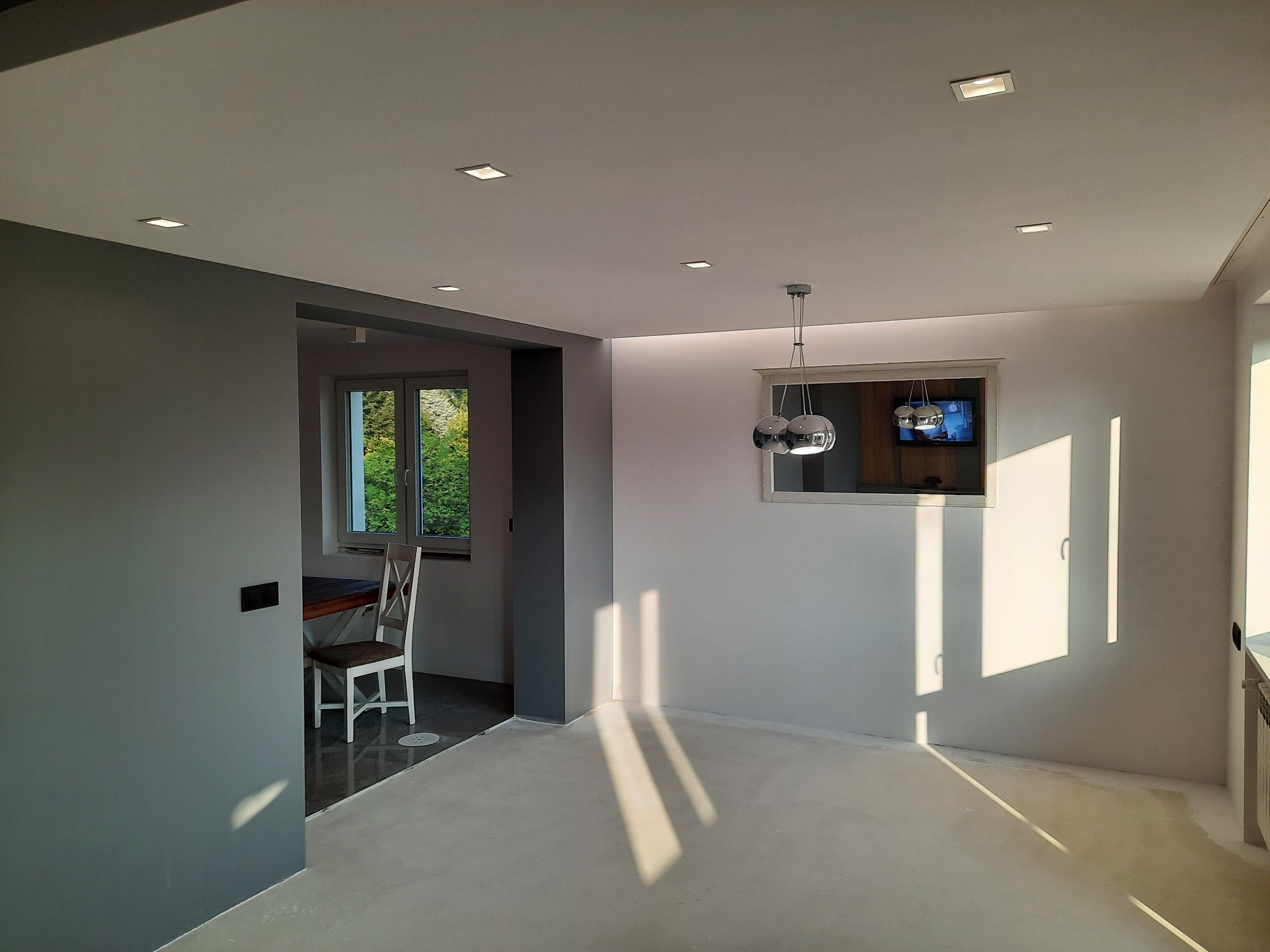 Nowoczesne wnętrze po remoncie, widok na pokój z szarą ścianą i jadalnią w tle, sufit z wbudowanymi punktowymi lampami, podłoga przygotowana pod wykończenie, wisząca lampa nad lustrem.