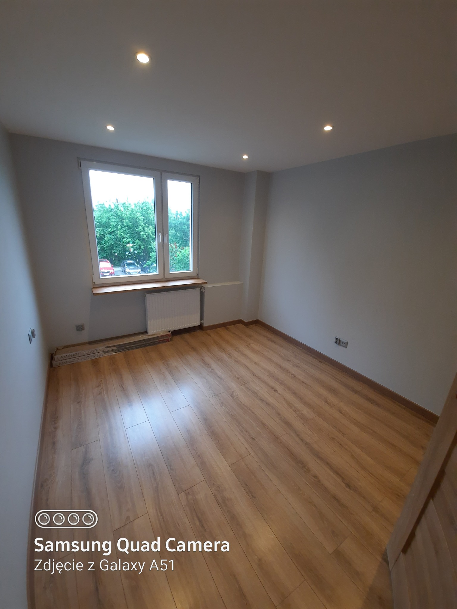 Pusty pokój po remoncie z panelami podłogowymi w odcieniach brązu, widok na okno z parapetem i grzejnikiem, sufit z punktowym oświetleniem.