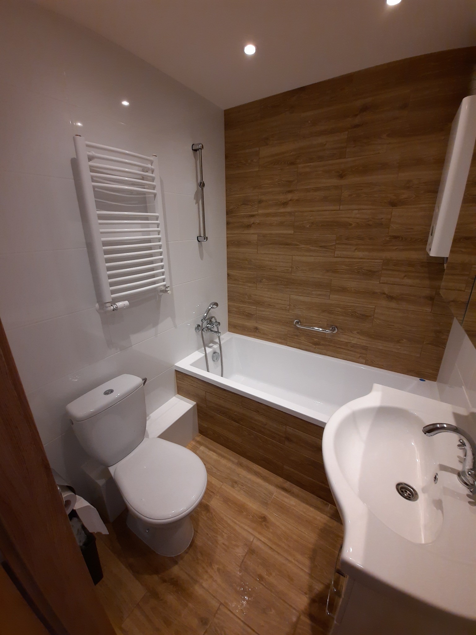 Wykończona łazienka z drewnopodobnymi płytkami na ścianie przy wannie, białą armaturą, grzejnikiem drabinkowym i podświetlanym sufitem.