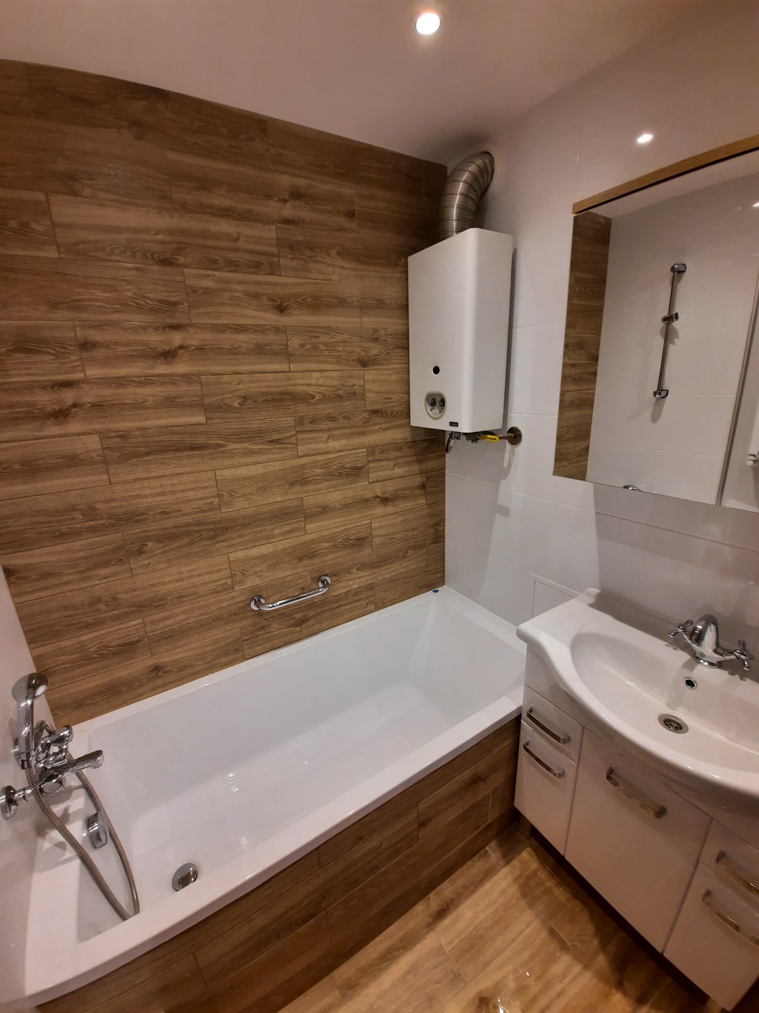 Wykończona łazienka z wanną obudowaną płytkami imitującymi drewno, białą umywalką z szafką i podgrzewaczem wody na ścianie.