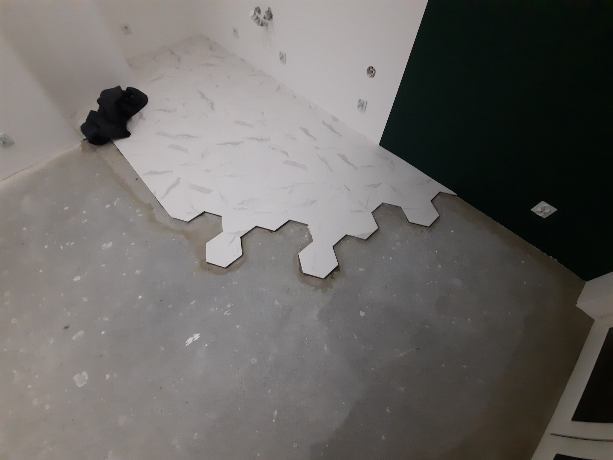 Układanie heksagonalnych płytek imitujących marmur na ścianie w trakcie prac wykończeniowych, widoczne niedociągnięcia i zabrudzenia na podłodze.