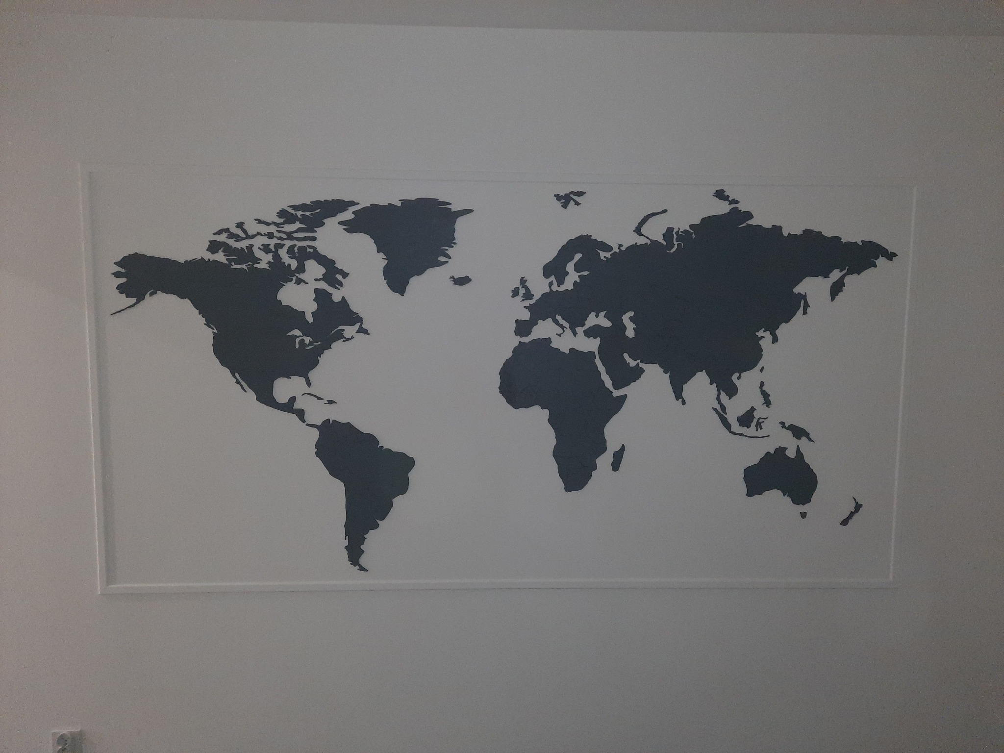 Grafika na ścianie przedstawiająca mapę świata w odcieniach szarości, umieszczona w białej ramie.